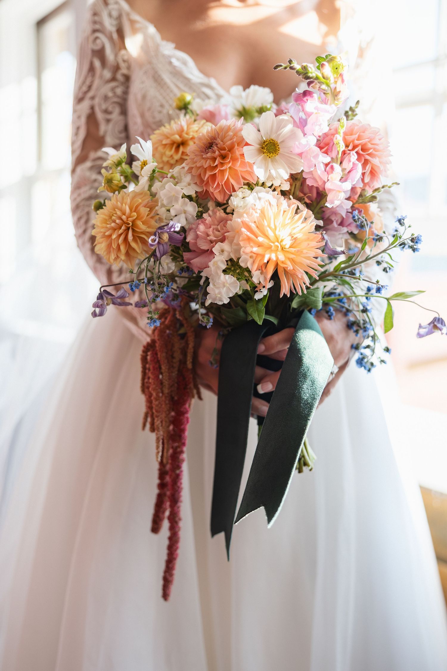Peach and pink dahlia bouquet with blue accents against white wedding dress.