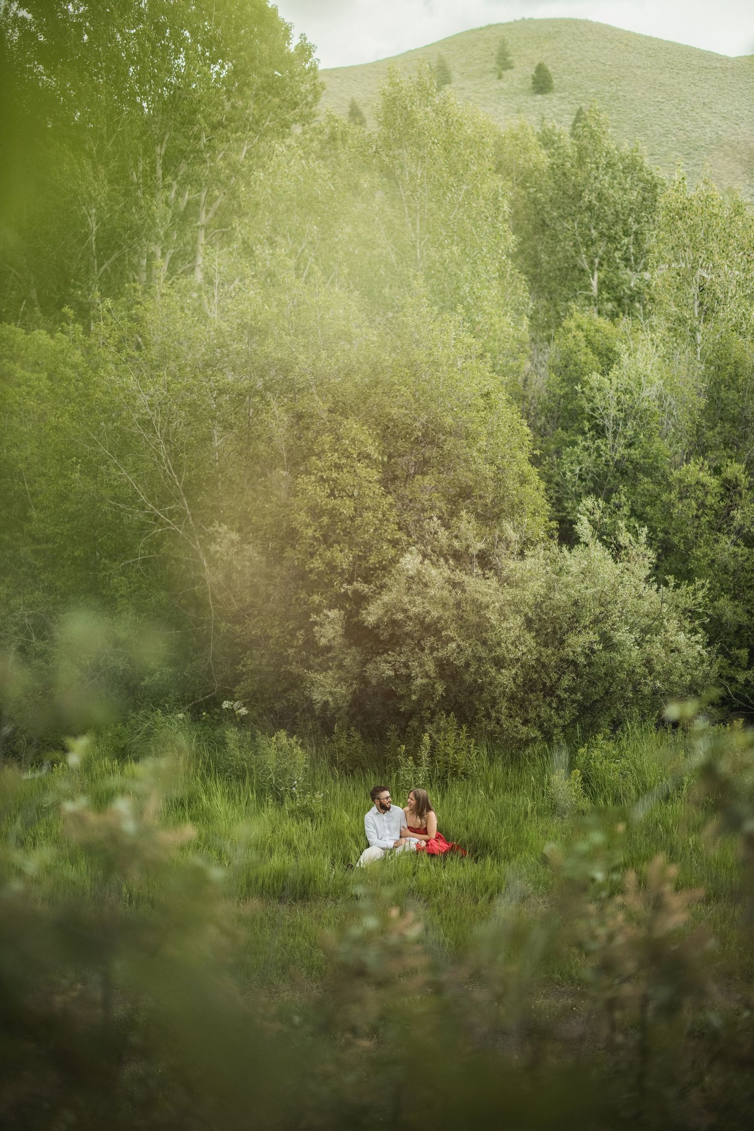 A couple sits together surrounded by lush green foliage and trees in a natural outdoor setting.