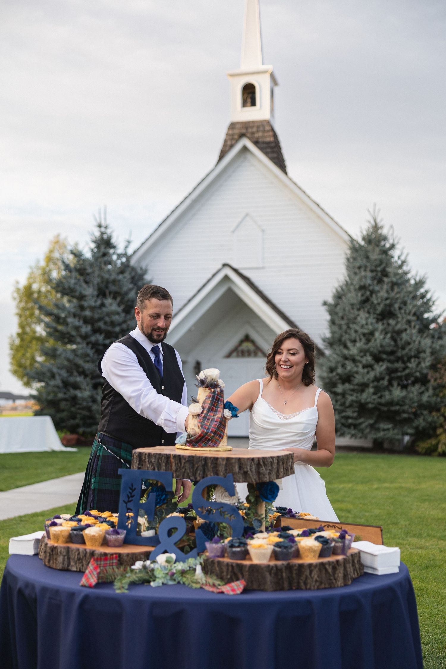A couple cuts their wedding cake on a rustic tiered display with a white chapel in the background.