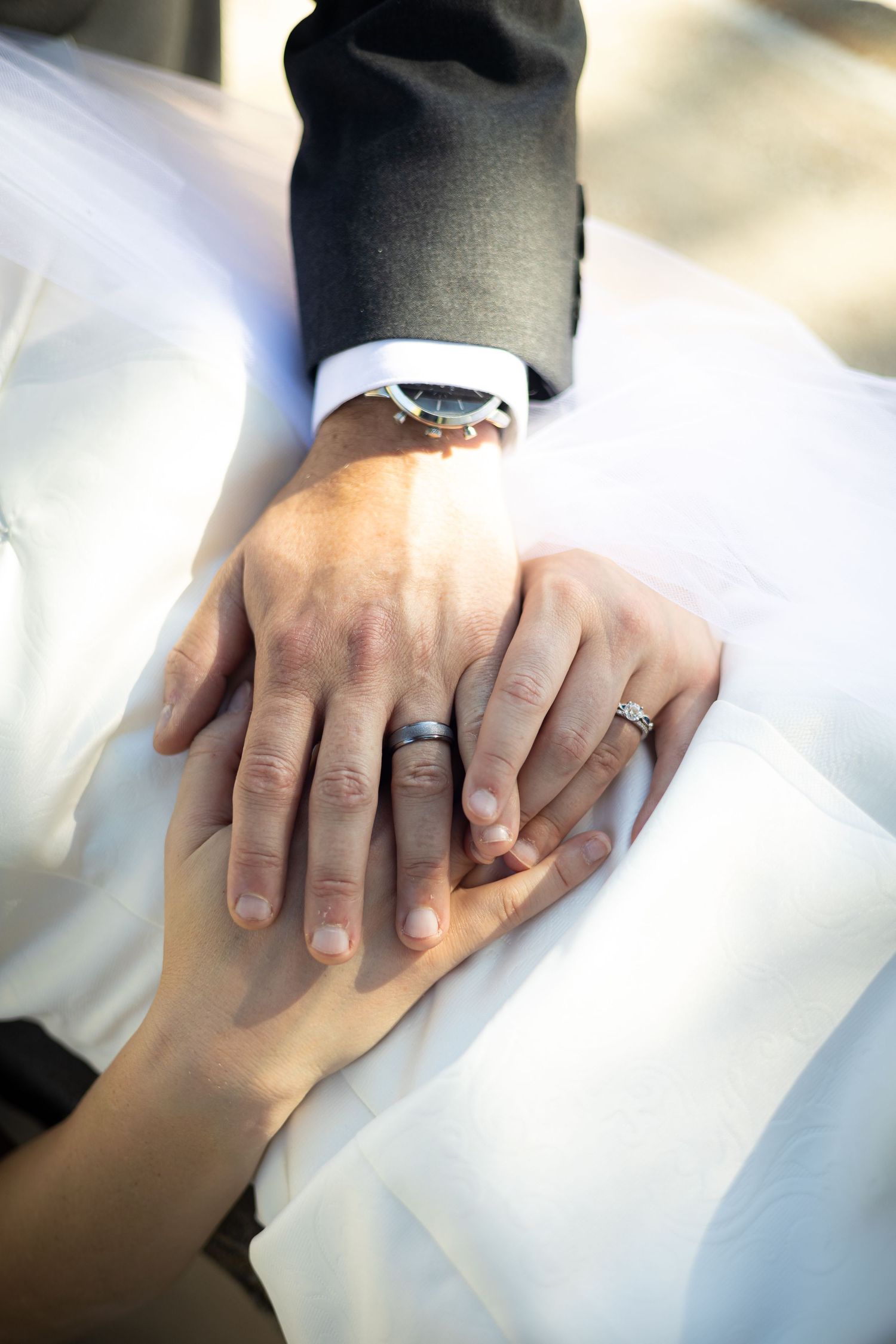 Close-up of wedding rings and hands clasped together on white dress fabric.