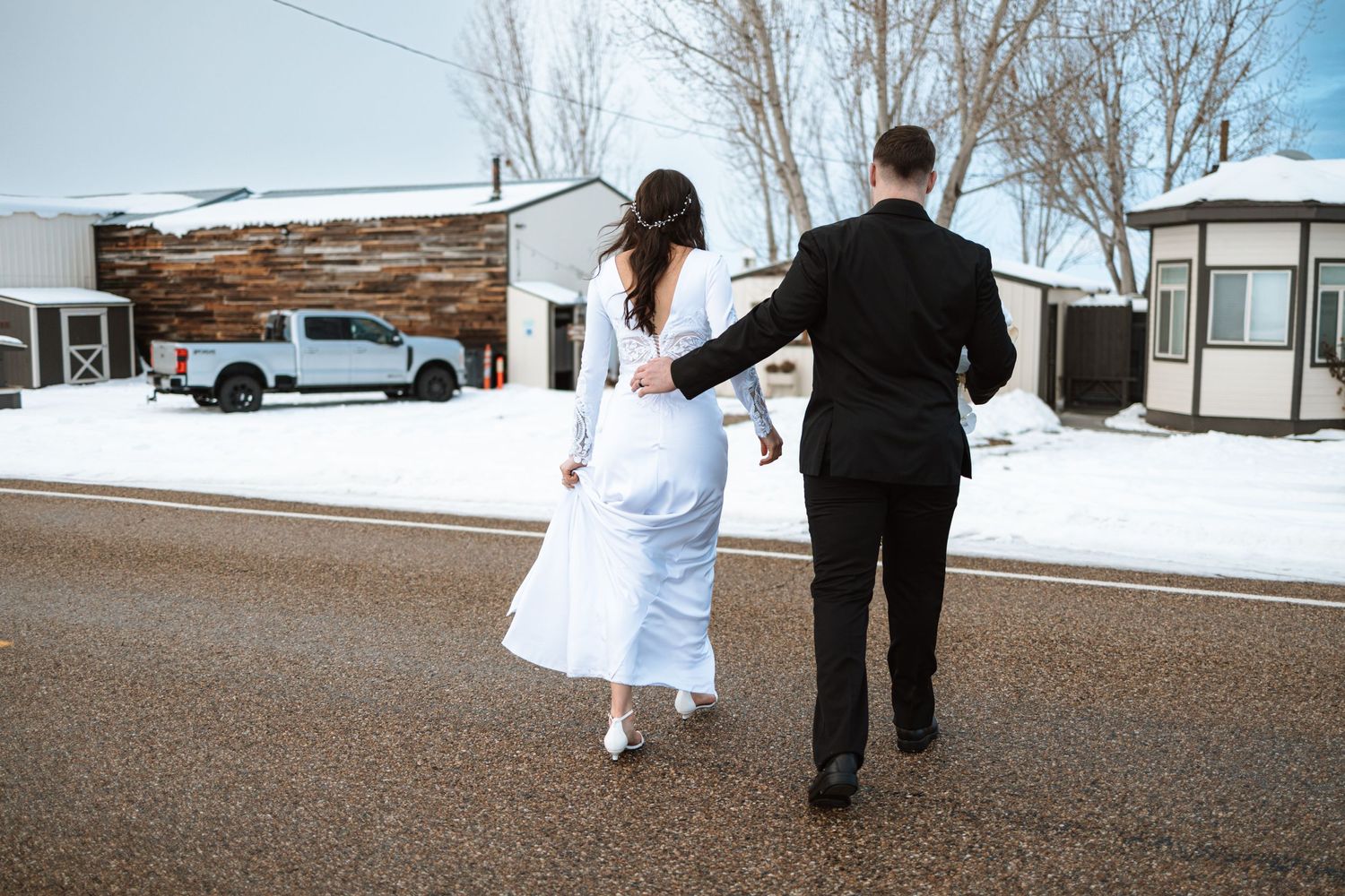 A couple walks away from camera on a snowy street in formal wedding attire.