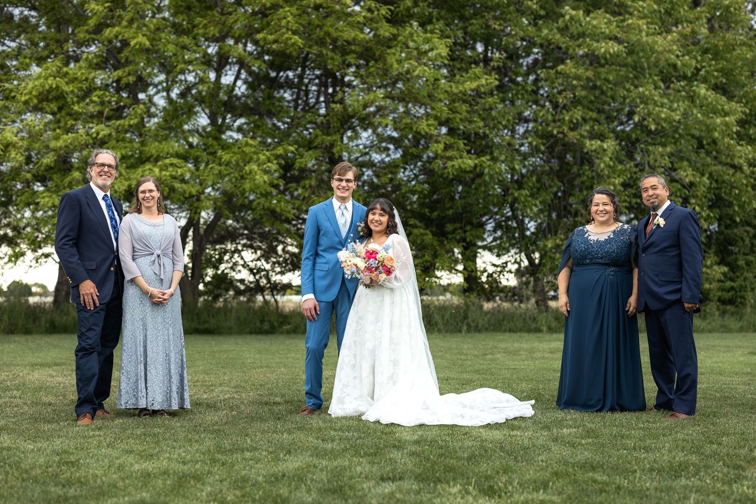 Wedding party poses for formal portrait on grass with large trees in background.