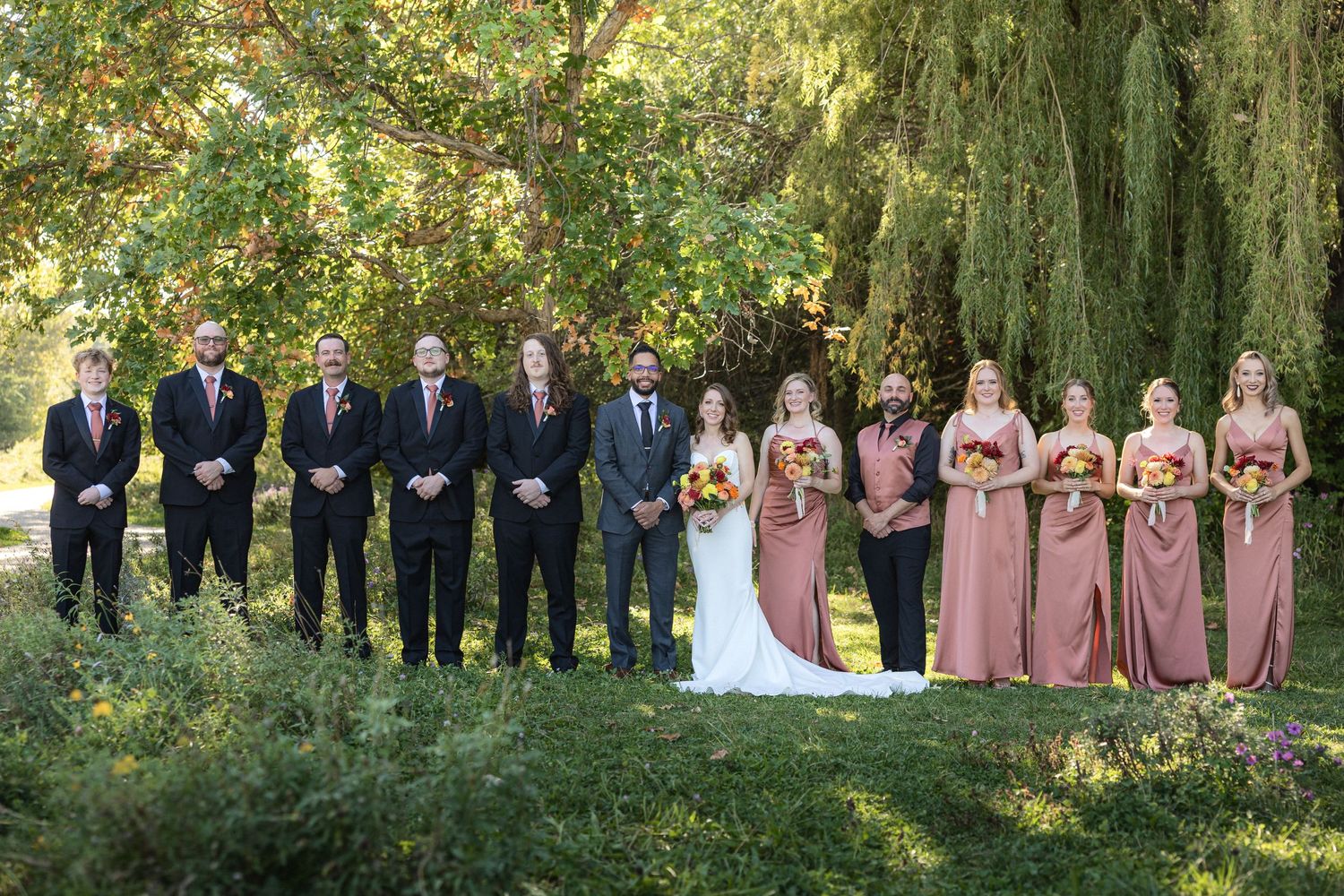 Wedding party poses outdoors with groomsmen in black suits and bridesmaids in blush pink dresses.