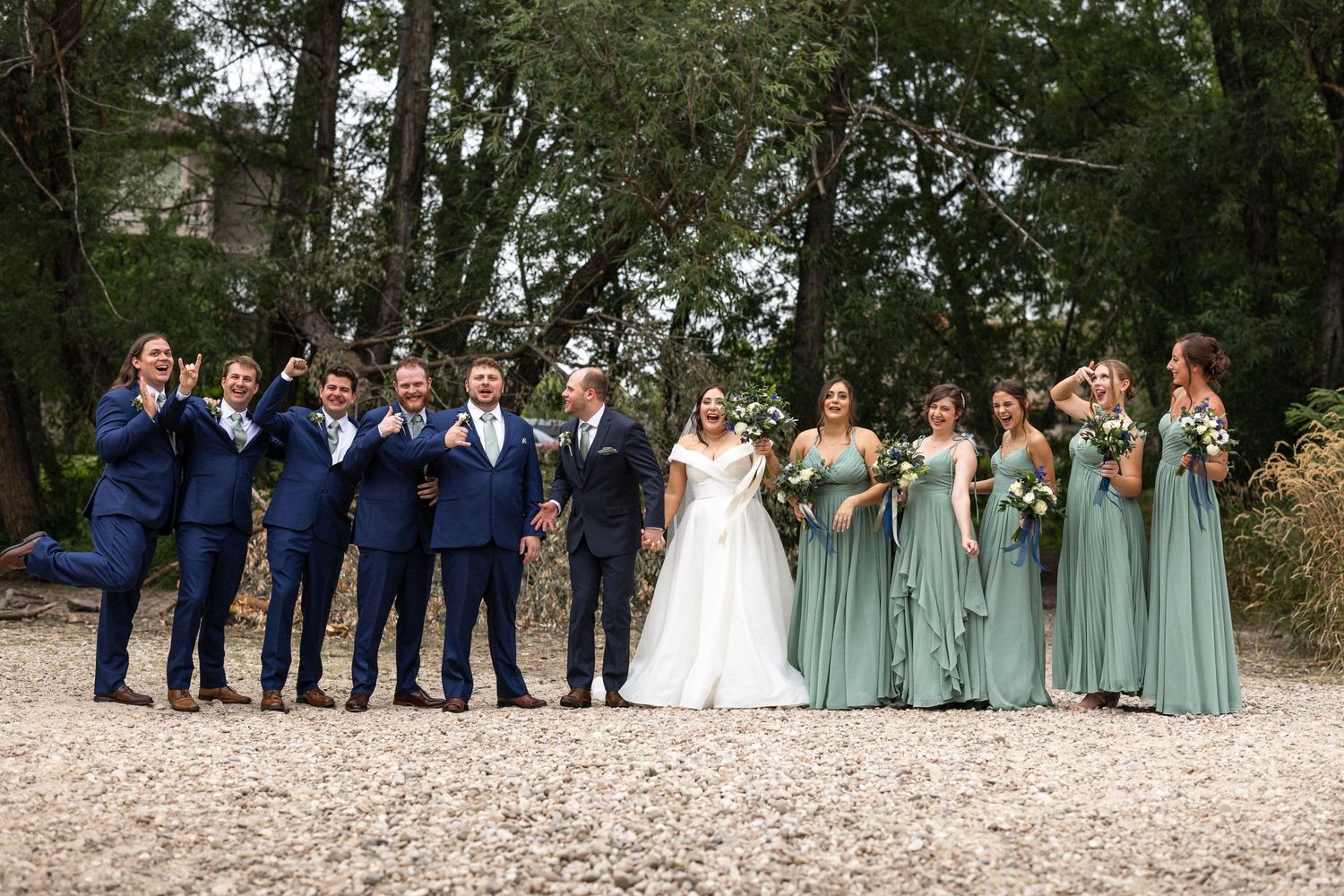 Wedding party poses outdoors with navy suits and sage green bridesmaid dresses against natural backdrop.