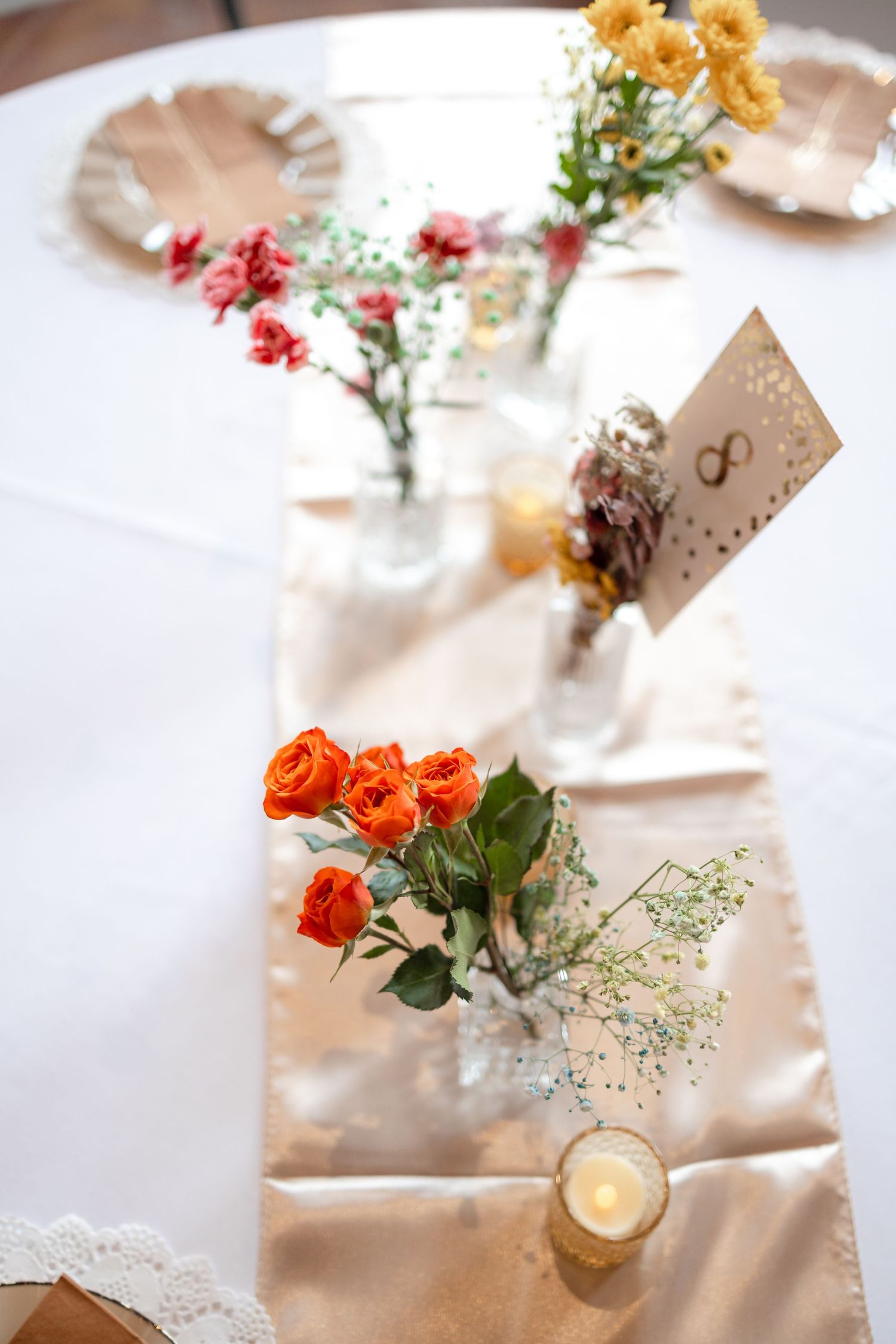 Wedding table setting with orange roses and wildflowers in small vases on white runner.