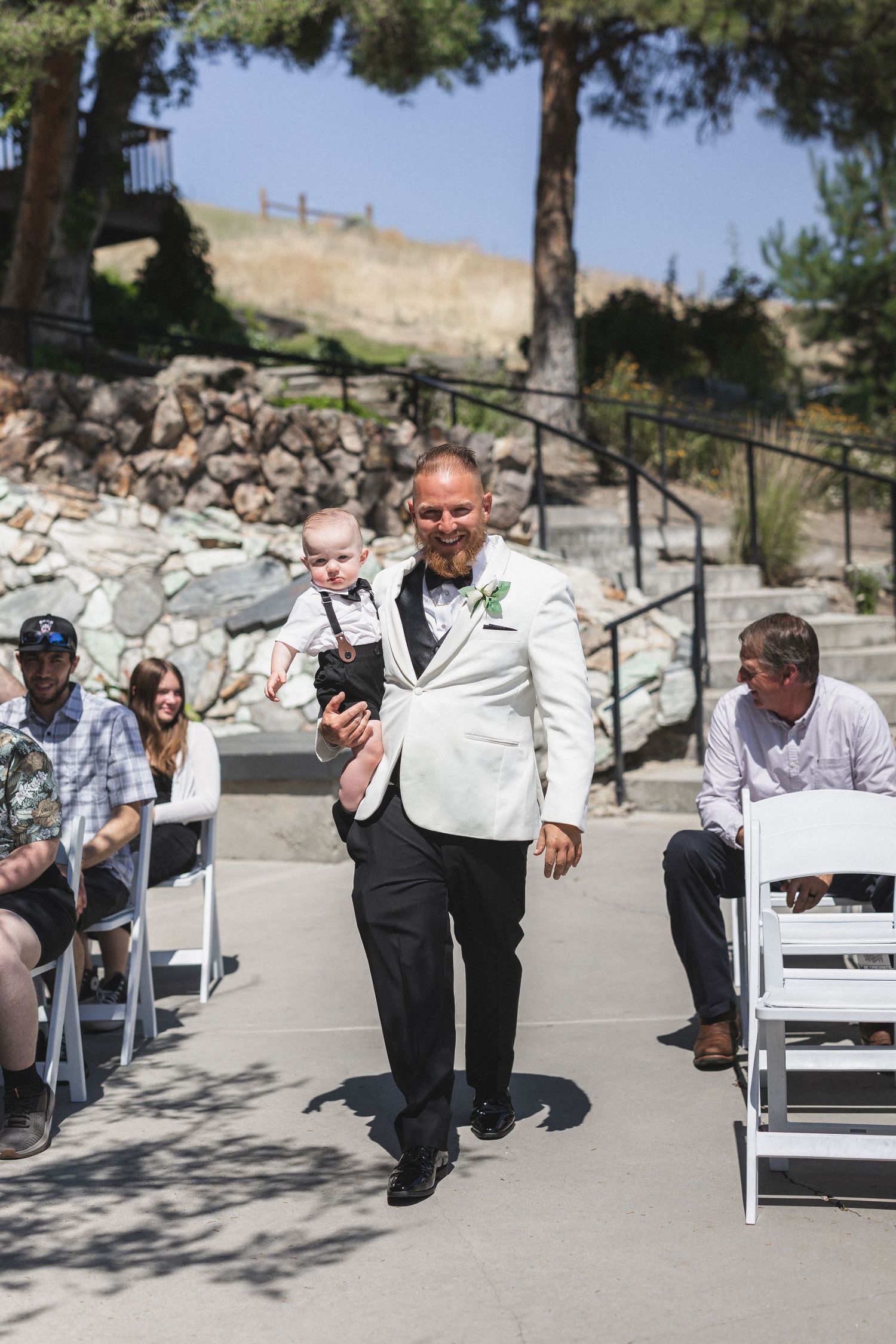 A person walks down the aisle carrying a baby at an outdoor wedding ceremony.