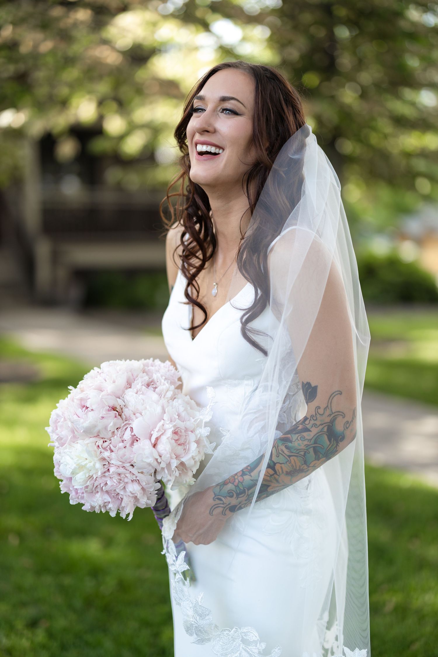Joyful bride laughing while holding pink peony bouquet in outdoor garden setting.