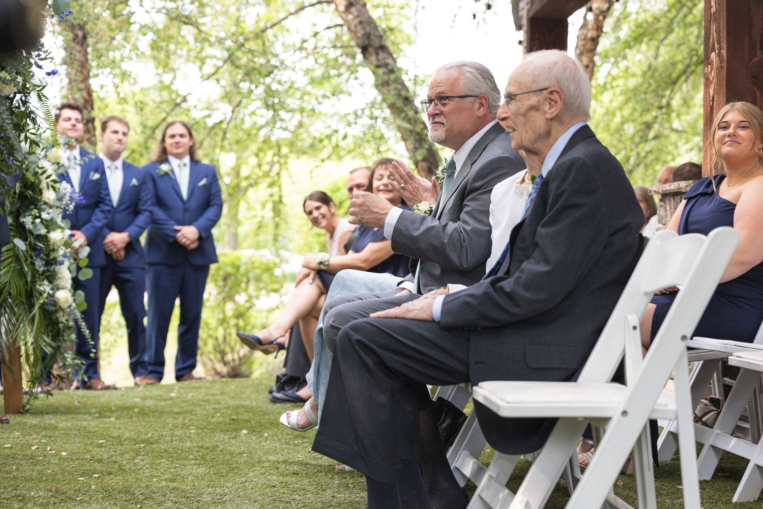 Wedding guests seated on white chairs watch an outdoor ceremony in a garden setting.