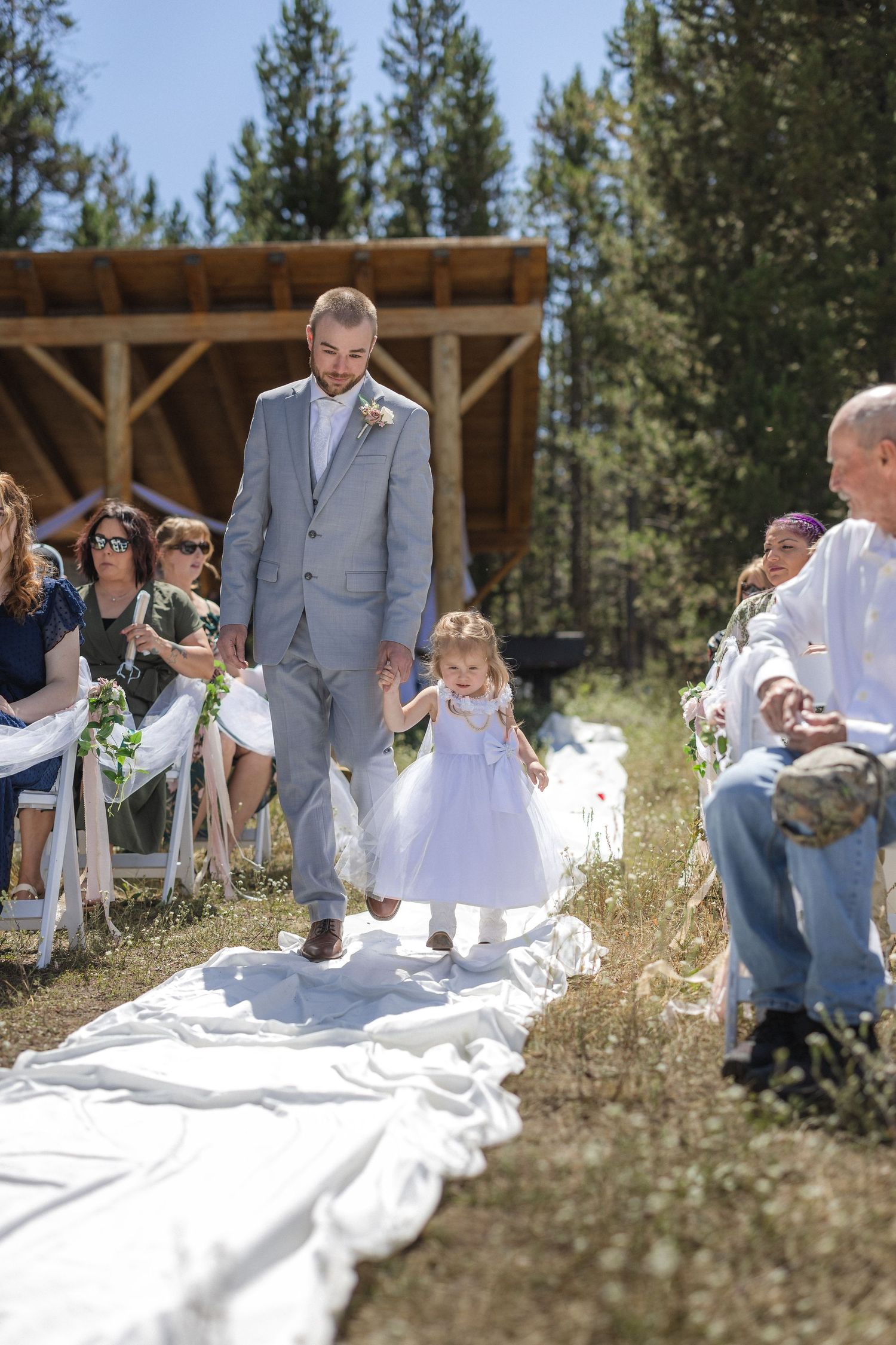 Wedding ceremony under wooden pavilion with person in grey suit and small child walking down white aisle.