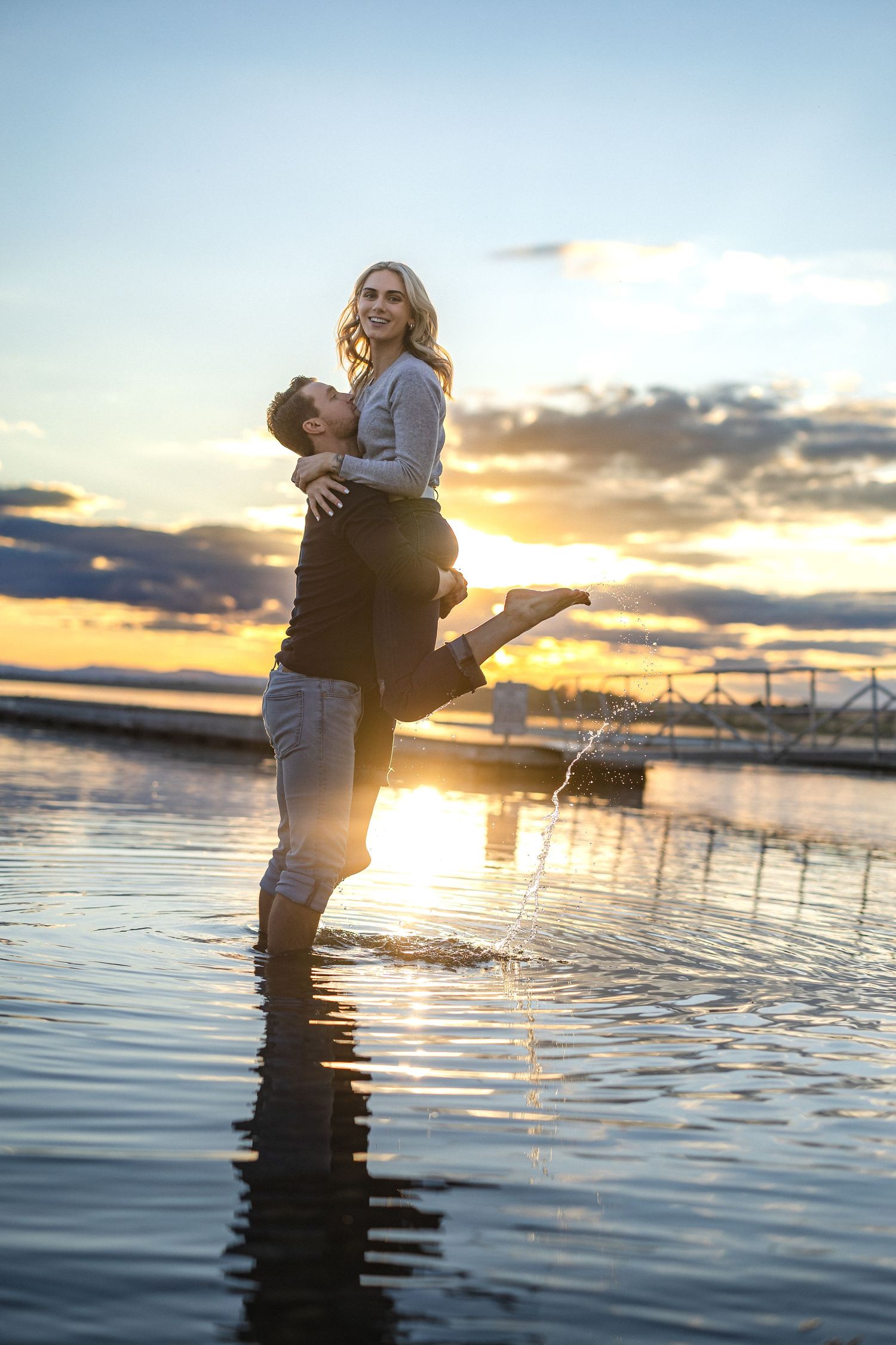 Couple standing in water during sunset with romantic lift pose near dock.