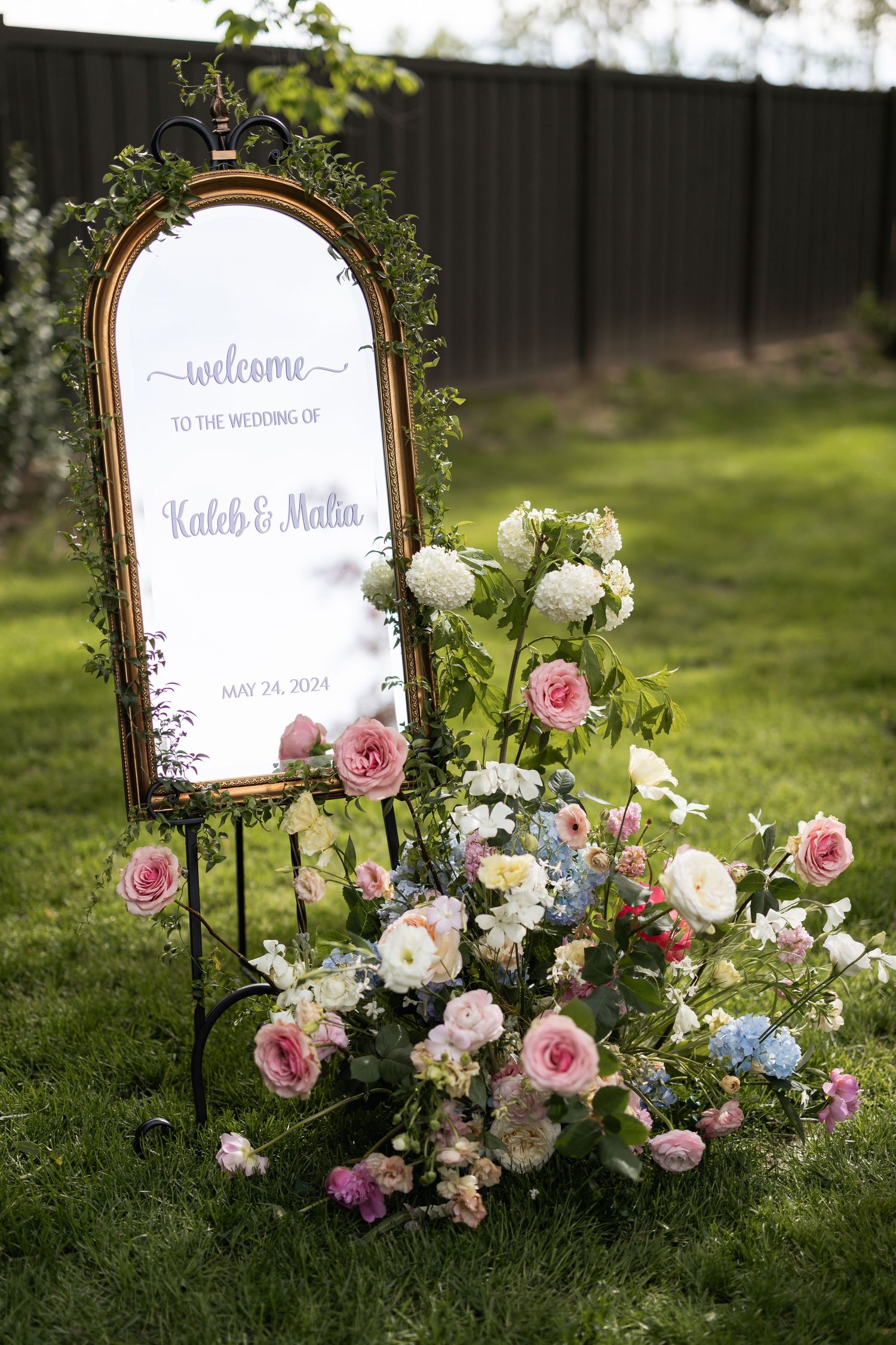 Vintage mirror decorated with pink and white roses displays a welcome sign in an outdoor garden setting.