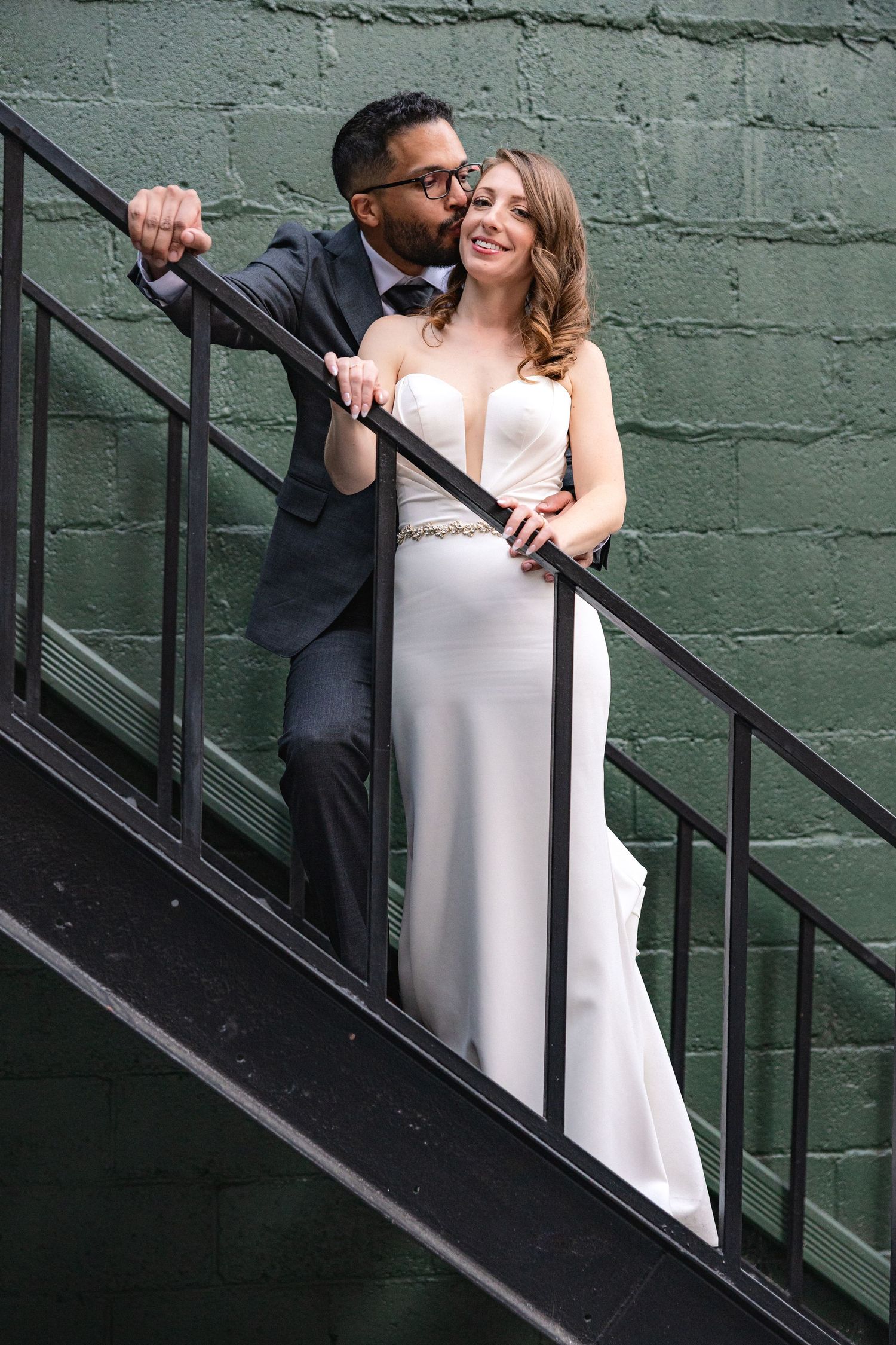 Wedding couple poses romantically on industrial metal staircase against green painted wall.