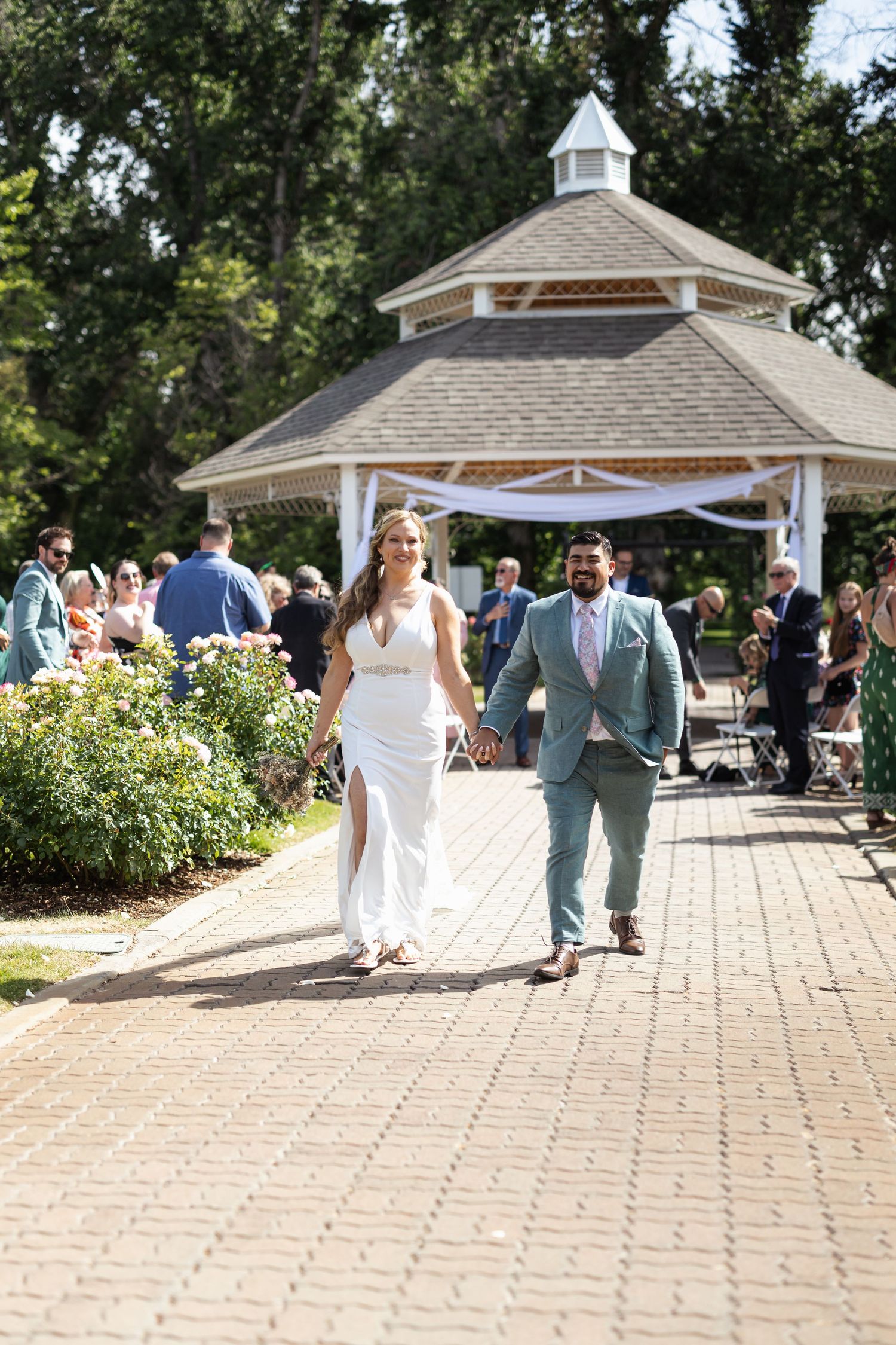 Newlyweds walk down brick pathway after ceremony at garden gazebo.