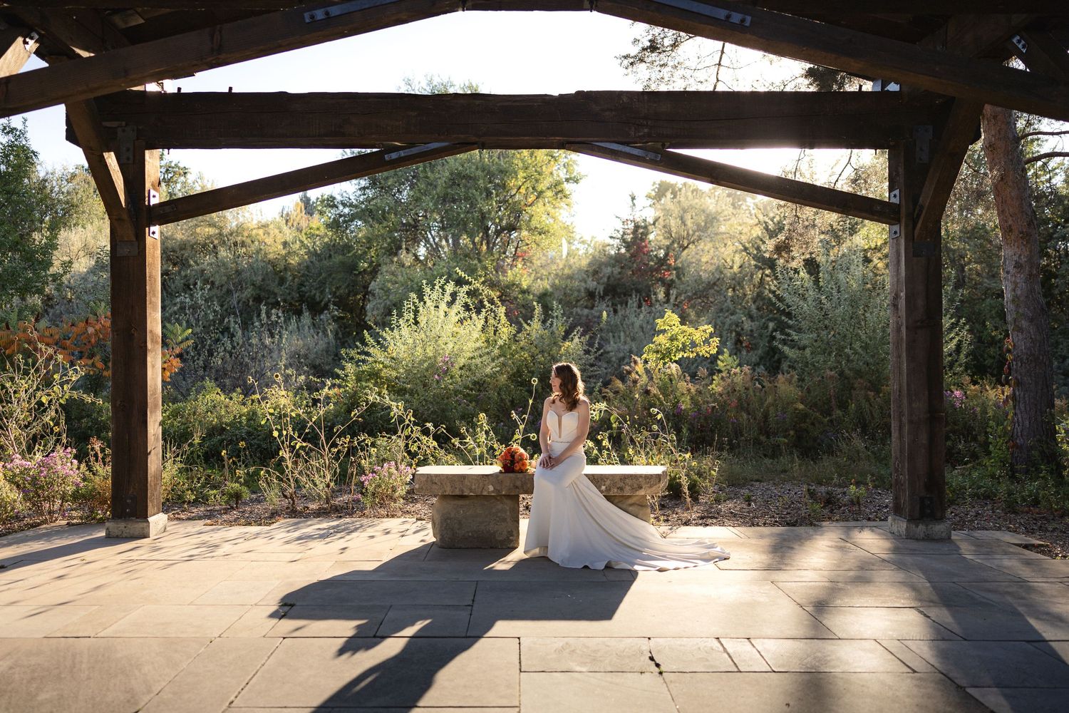 A wedding couple poses under a rustic wooden pavilion surrounded by lush garden landscaping at sunset.