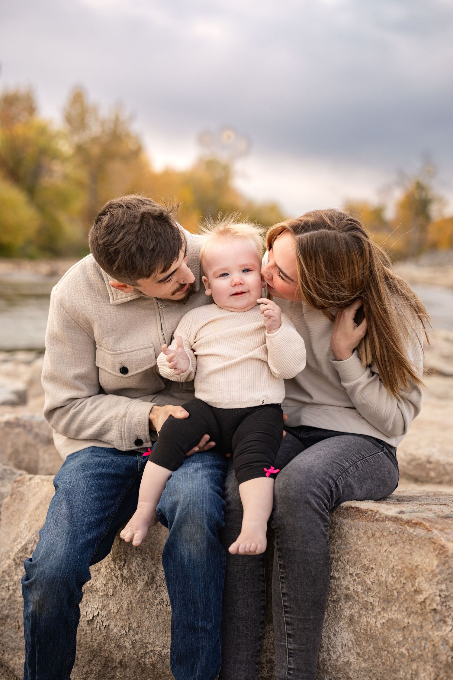 A family shares a tender moment together while sitting on rocks during a fall afternoon with autumn foliage in background.