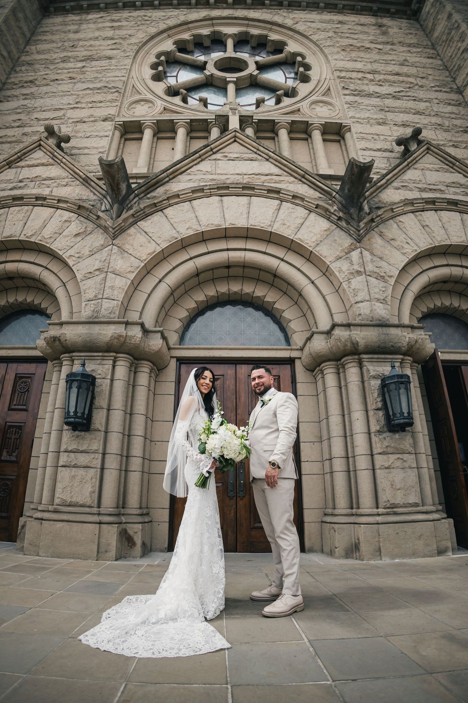 Bride and groom pose in front of a grand stone church entrance, bride holding white bouquet with long lace veil train.