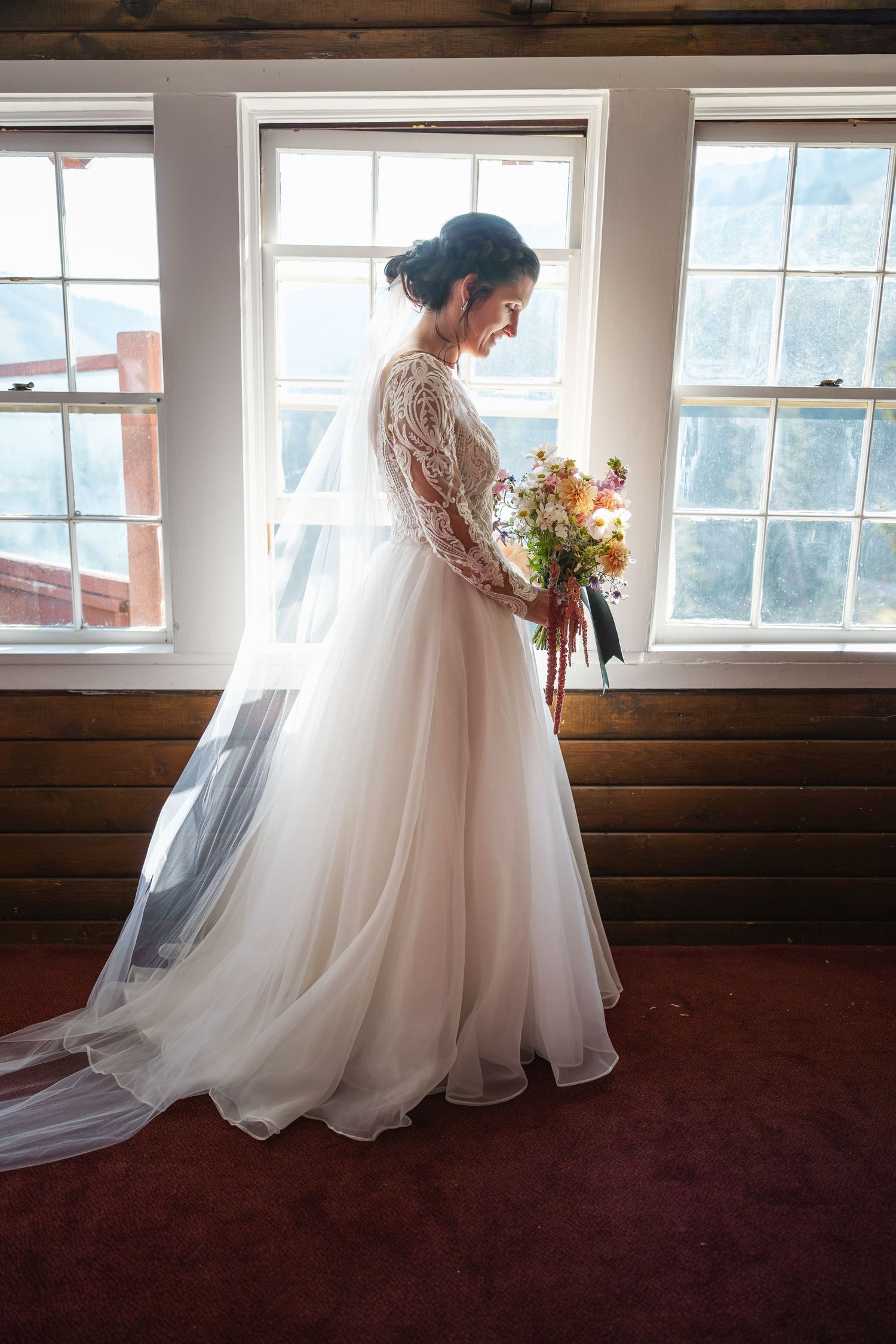 Flowing white wedding dress with lace details silhouetted against large windows in a sunlit room.