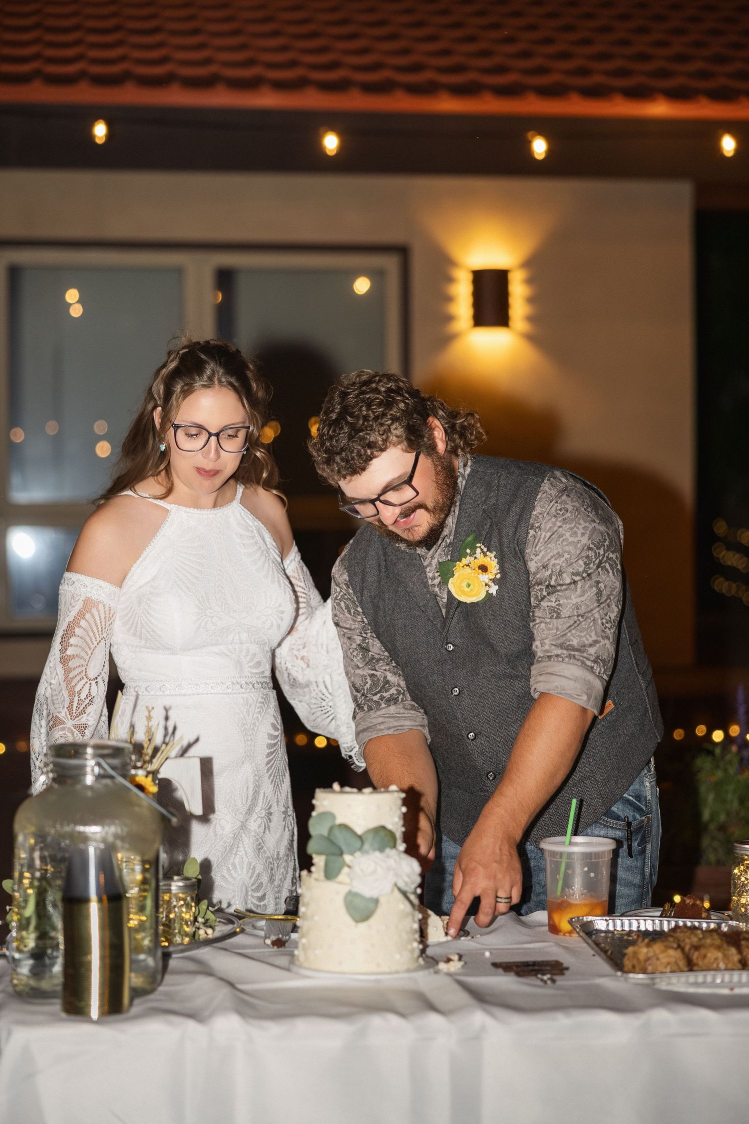 Wedding reception dessert table with string lights and people cutting cake together.