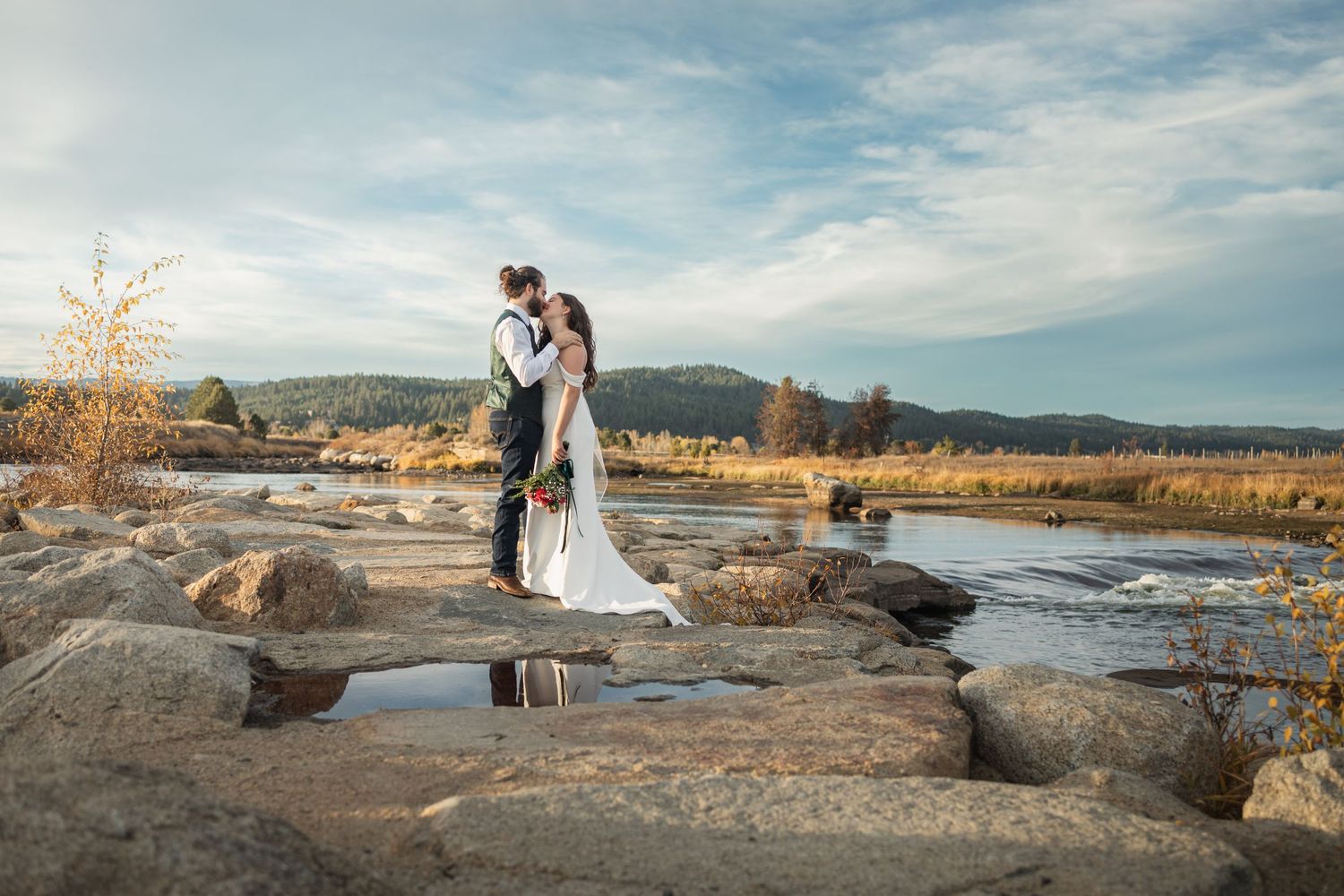 Newlyweds standing on natural rock formation near flowing river during outdoor wedding photoshoot.