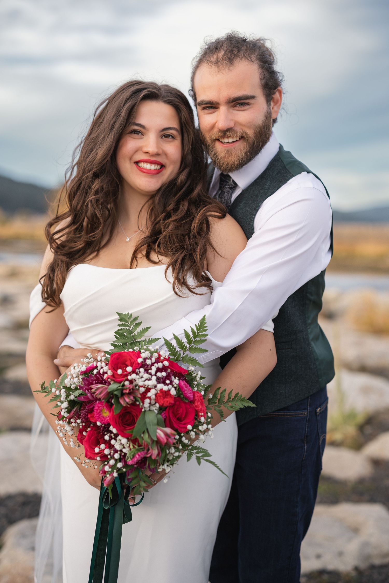 Joyful wedding portrait with red floral bouquet against rugged outdoor landscape.