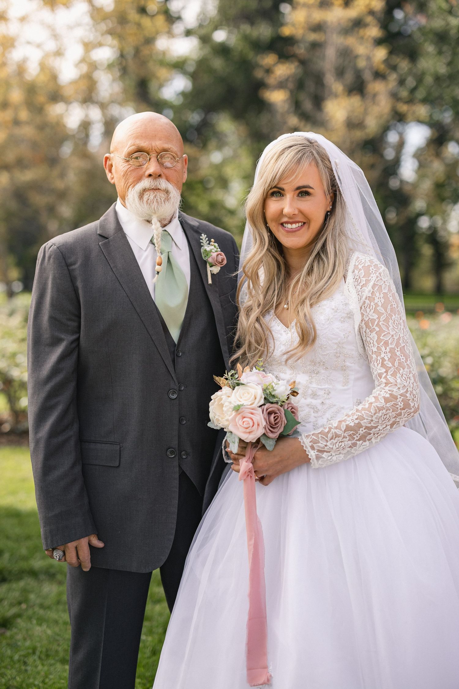 A bride in a lace wedding gown stands with an older gentleman in a gray suit during an outdoor garden ceremony.
