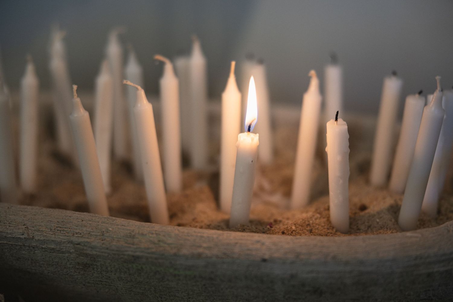 A collection of white candles arranged in sand with one flame burning brightly.