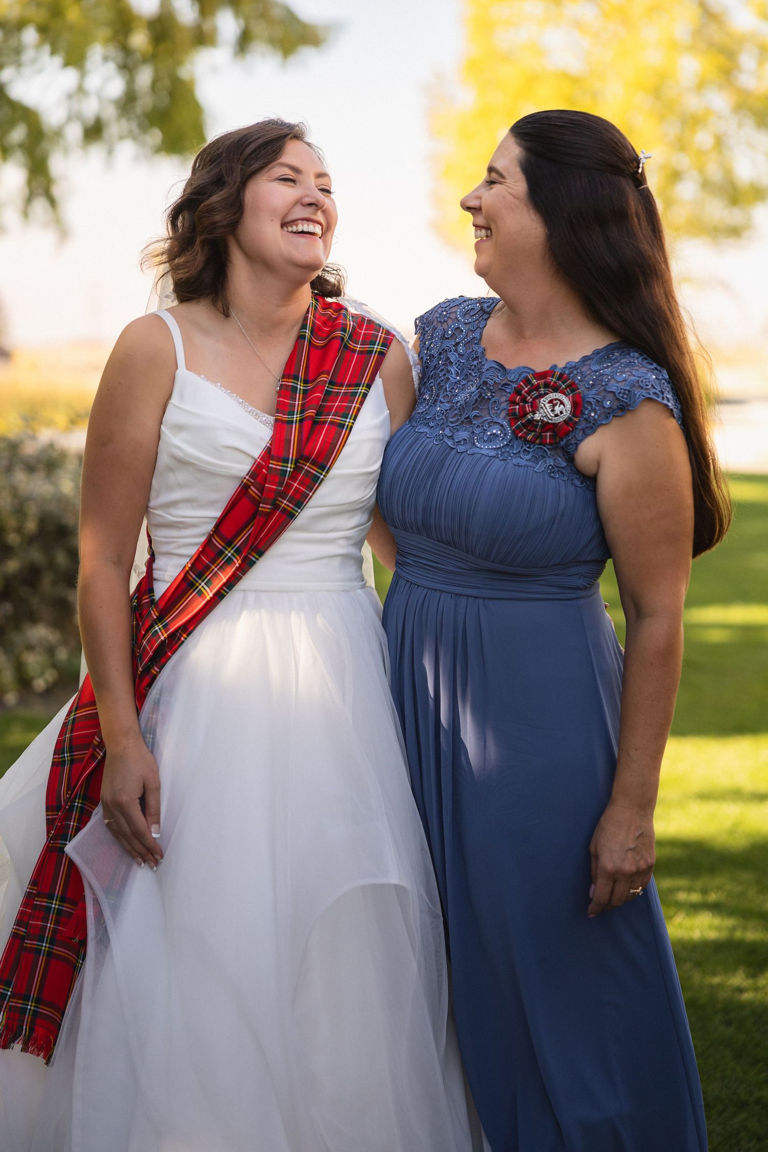A bride in white wearing a tartan sash shares a joyful laugh with someone in a navy blue dress outdoors.