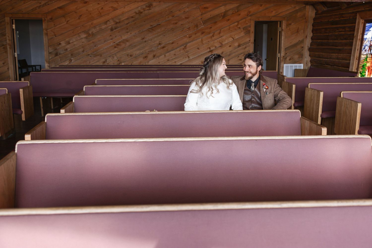 Rustic chapel interior with wooden beams and rows of burgundy pews bathed in warm light.