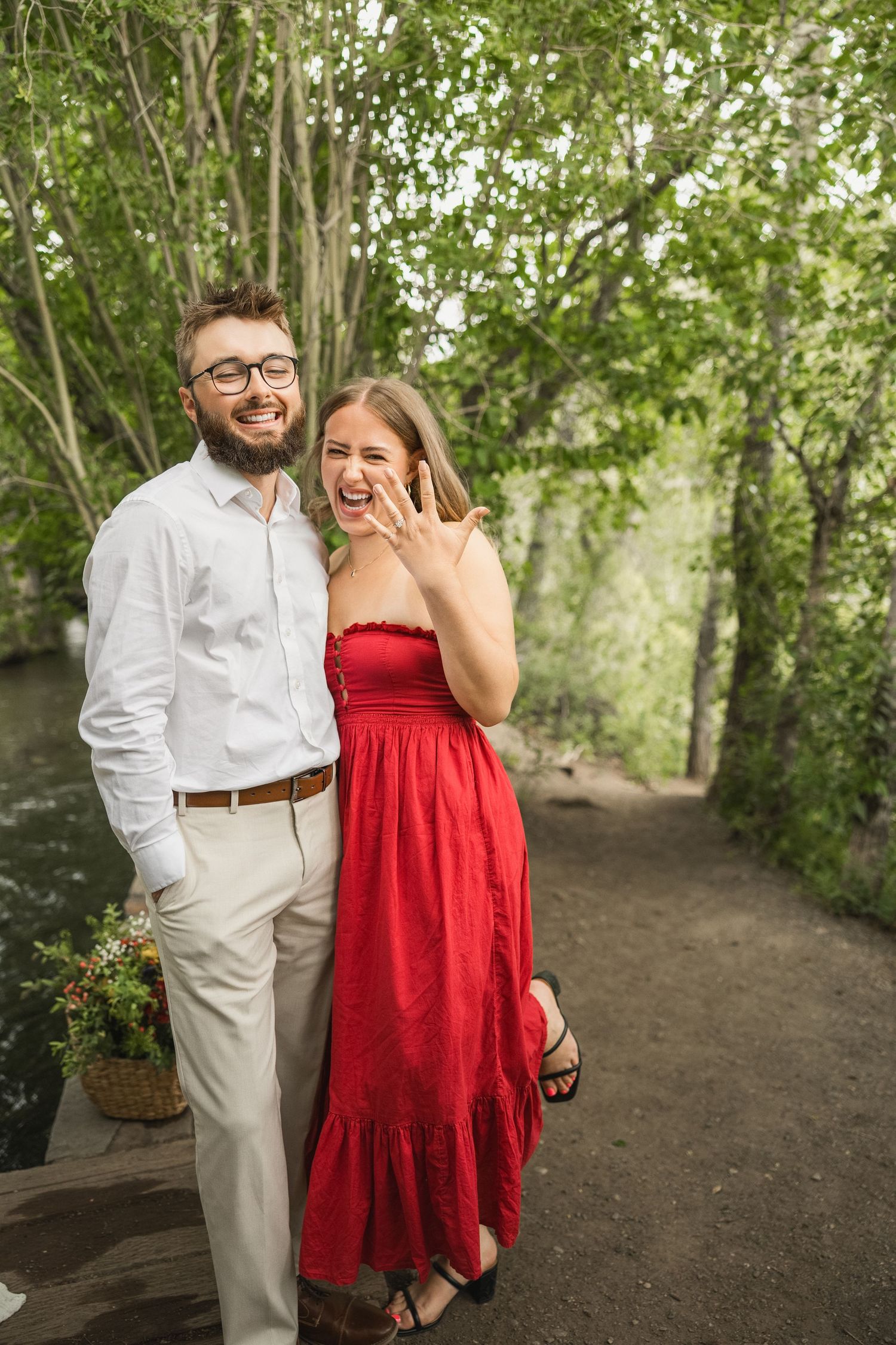 Couple celebrating engagement on woodland path with woman in red dress showing ring and joyful expressions.