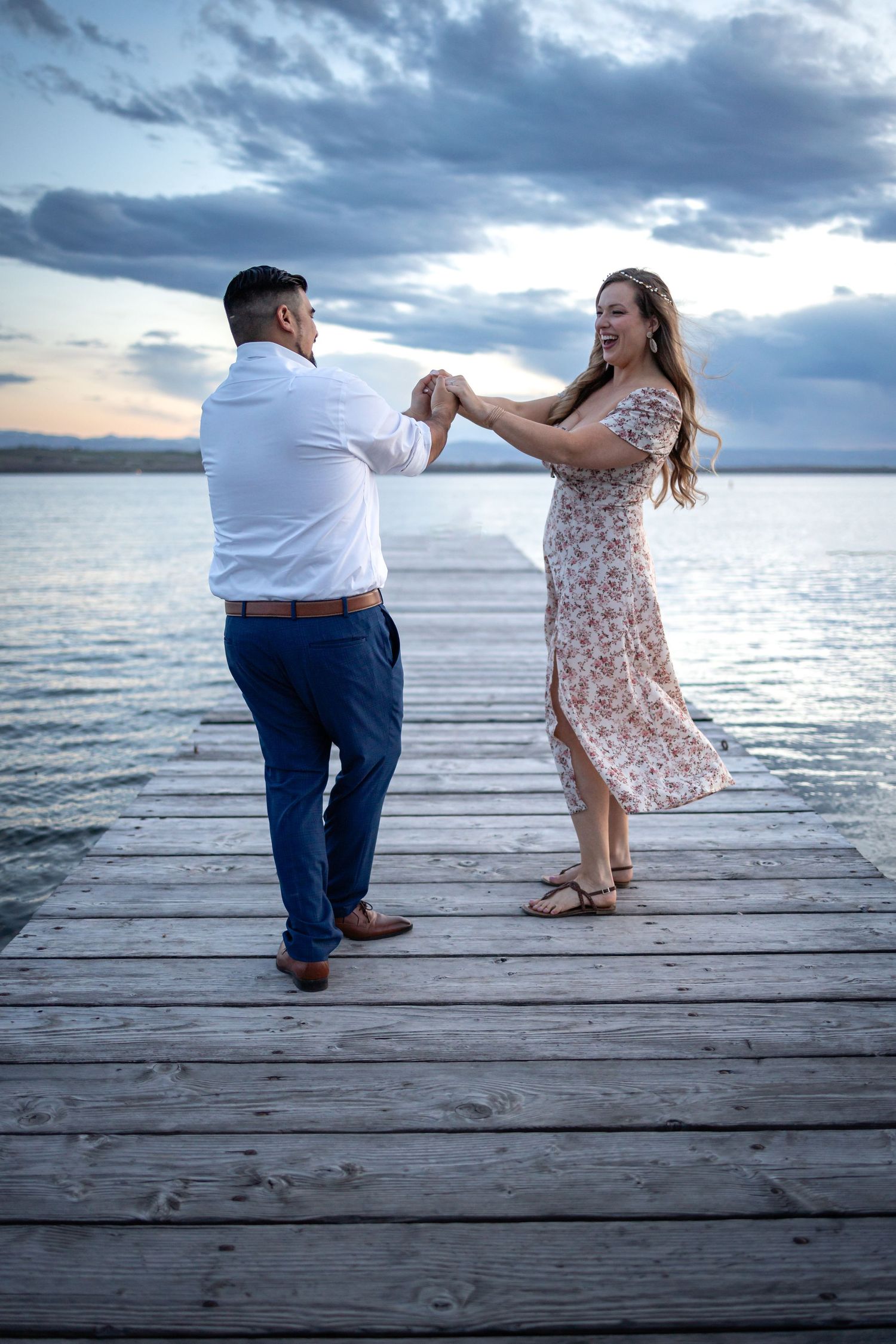 A couple dances romantically on a wooden pier at sunset with dramatic cloudy skies over a calm lake.