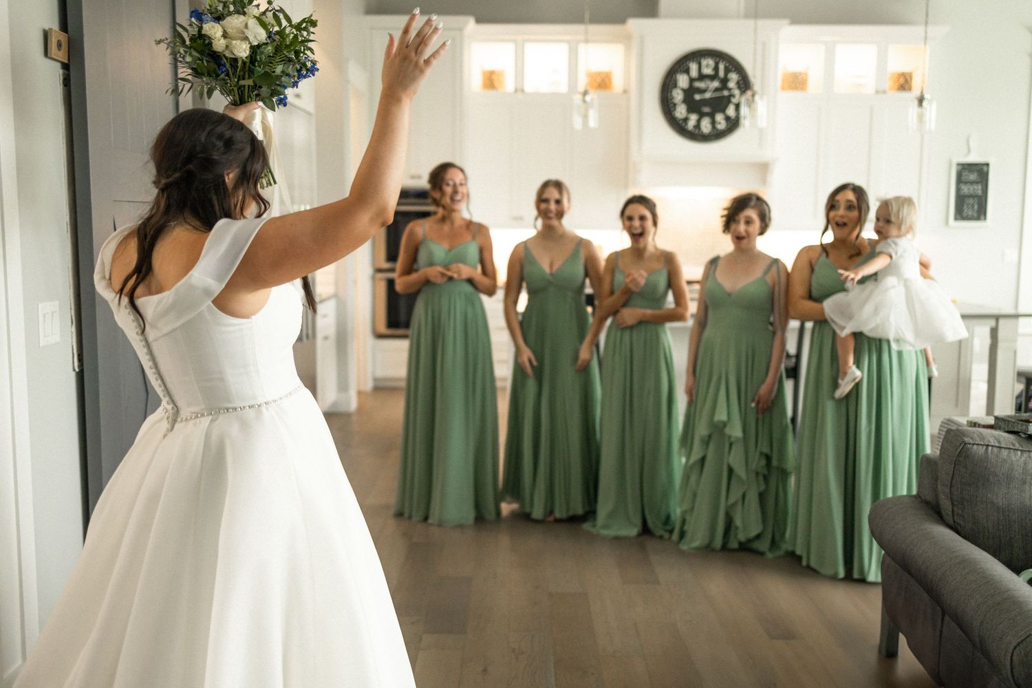 Group of bridesmaids in matching sage green dresses react during wedding preparation.