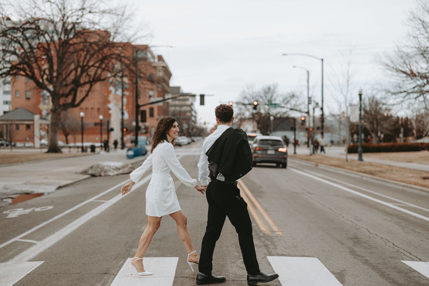 A playful couple in formal wear dances across an urban crosswalk.