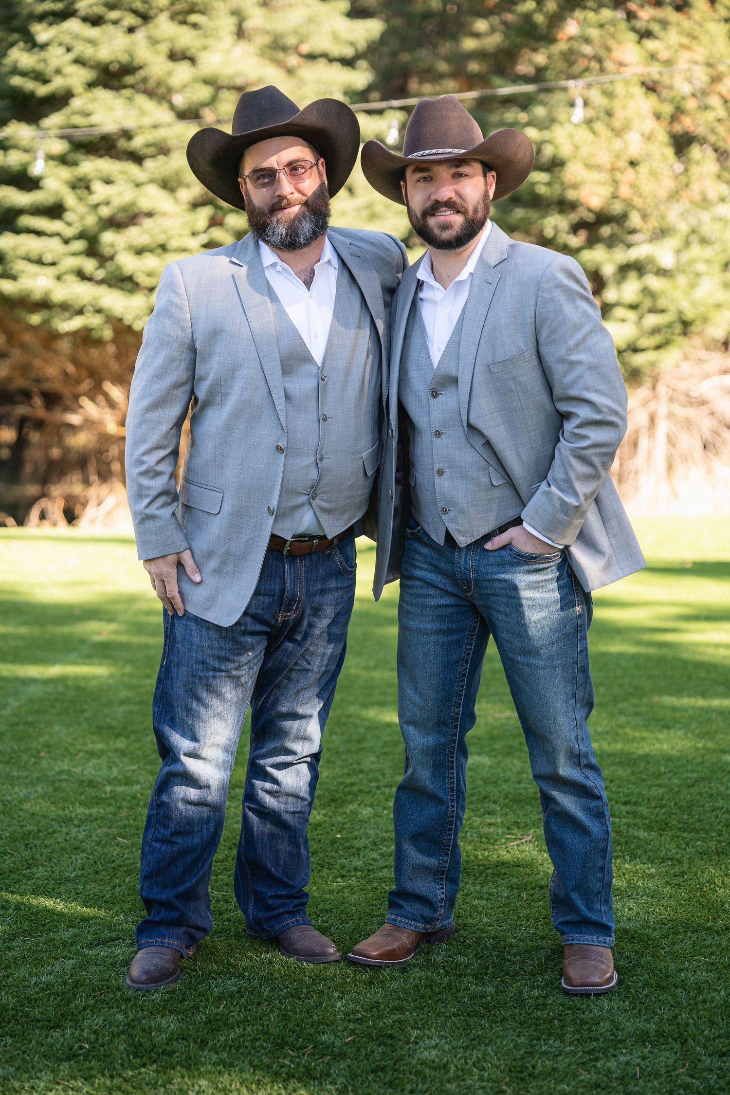 Two groomsmen in matching gray suits and cowboy hats pose together on grass.