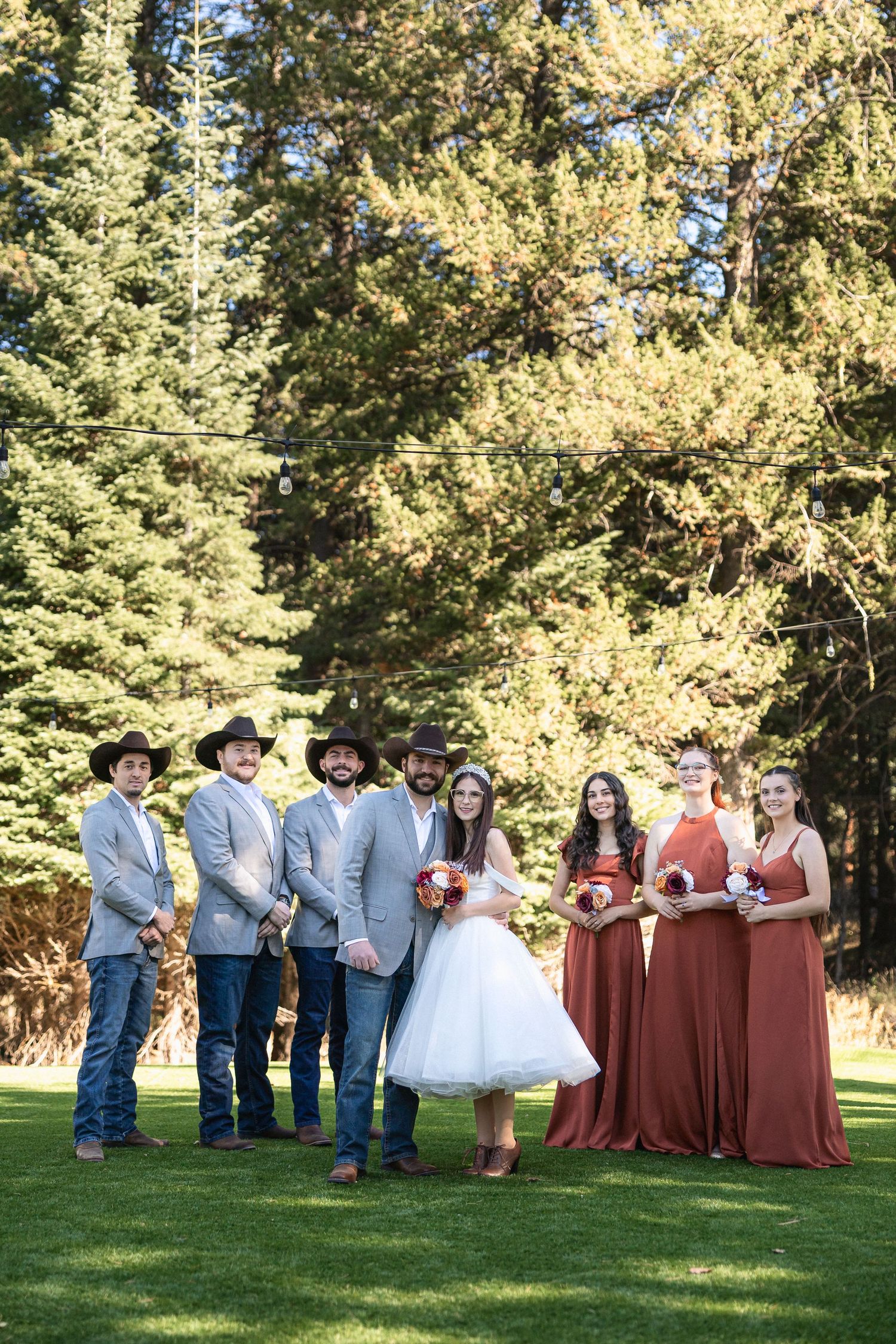 Rustic outdoor wedding party photo with groomsmen in cowboy attire and bridesmaids in terracotta dresses.