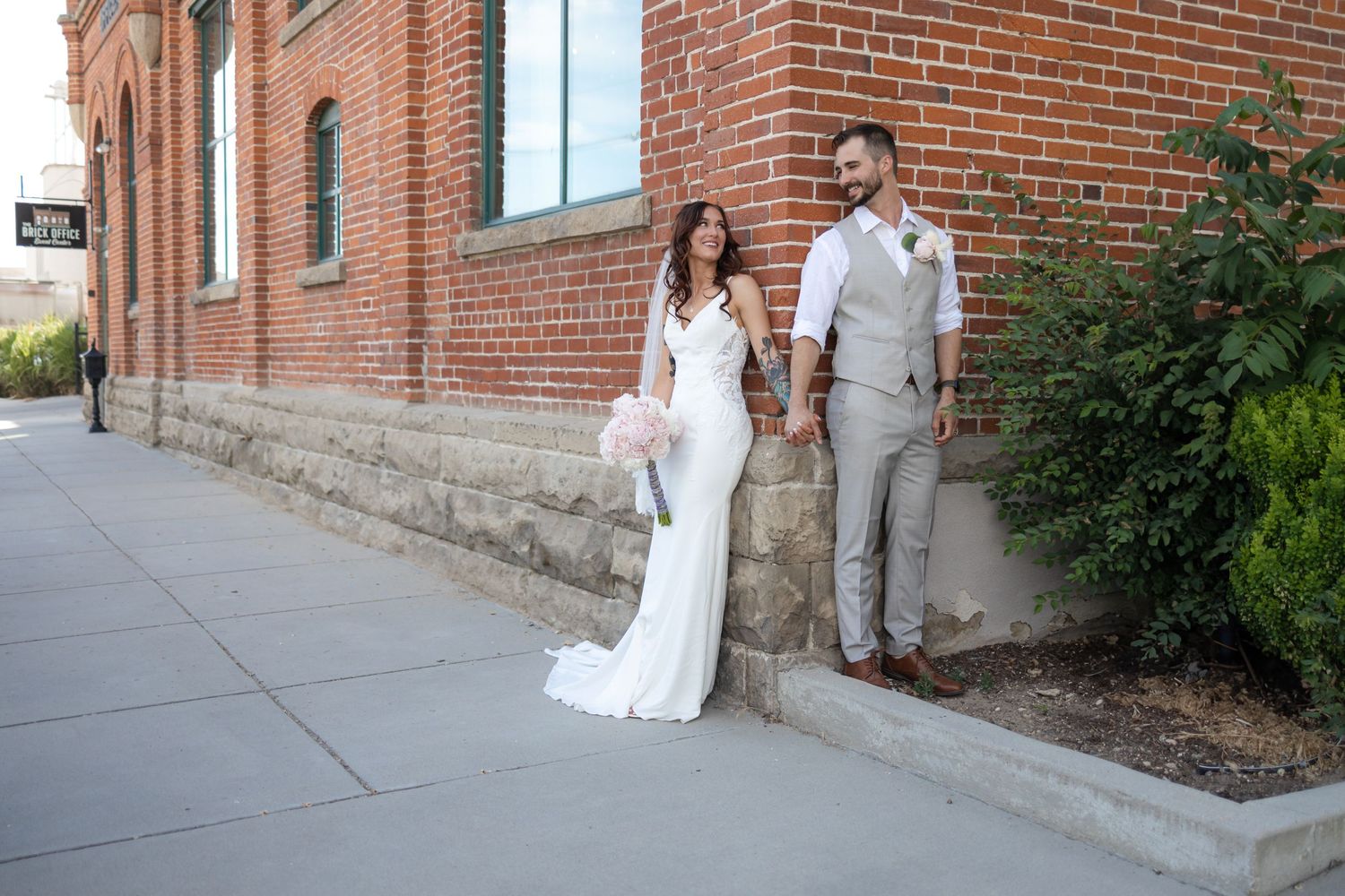 Elegant wedding couple poses against rustic brick wall exterior in white dress and light gray suit.
