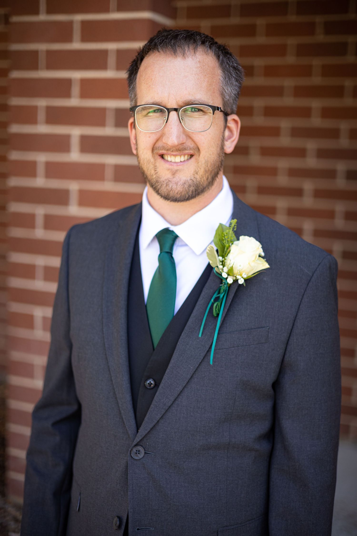 Man in formal dark suit with green tie smiles while wearing white rose boutonniere.