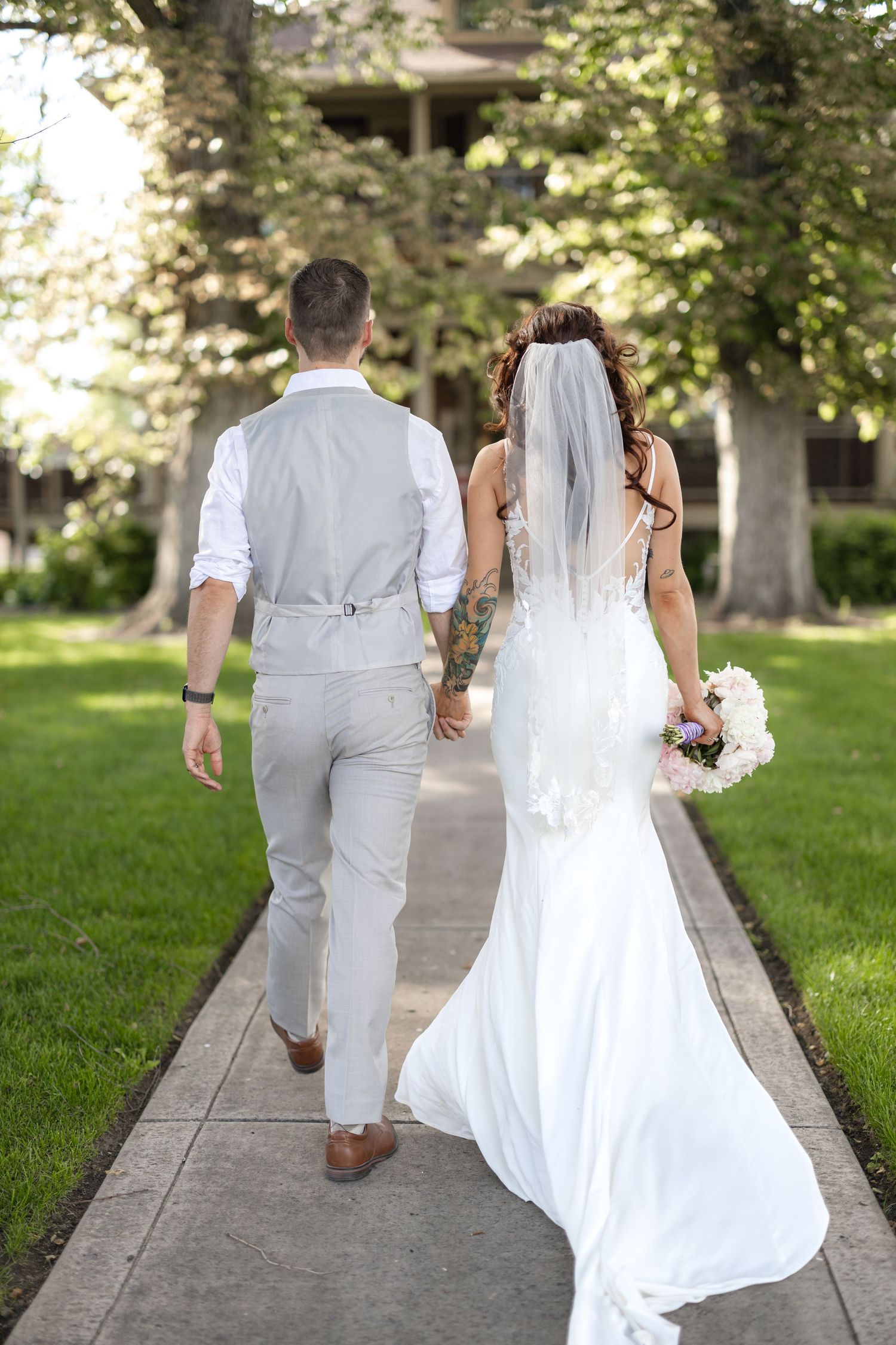 Newlywed couple walking down a tree-lined path, with the bride in a flowing white dress holding a bouquet.