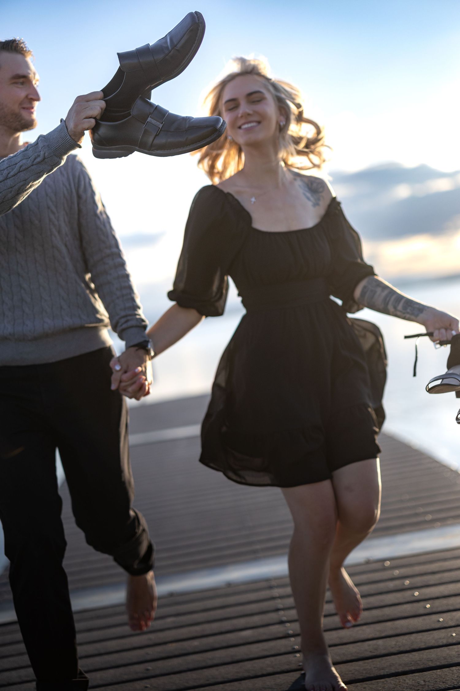Two people holding shoes and dancing playfully on a dock at sunset.
