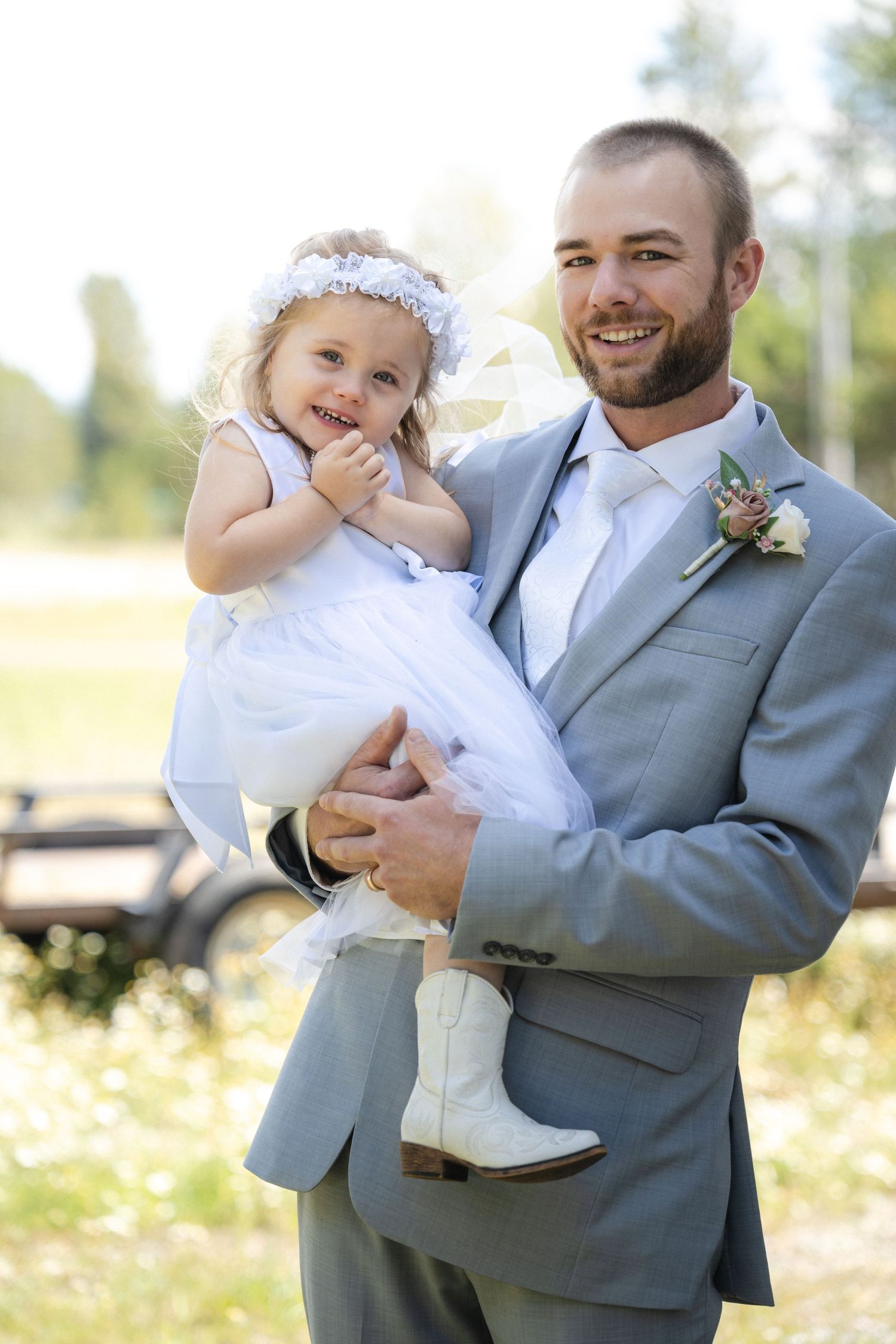 A groom in grey suit holds a smiling young flower girl wearing white dress and crown.