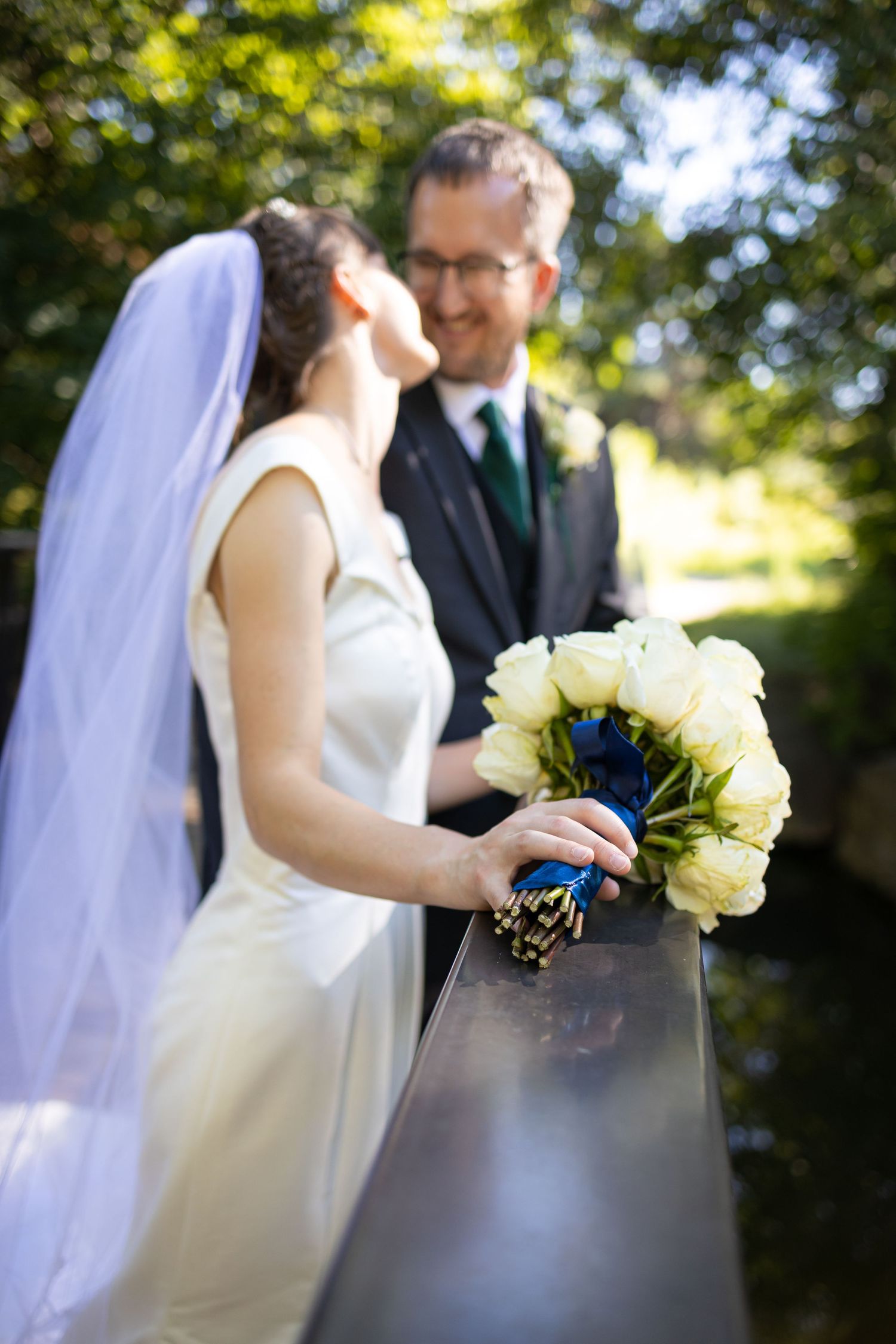 Wedding couple poses with white bouquet on a sunny outdoor walkway.
