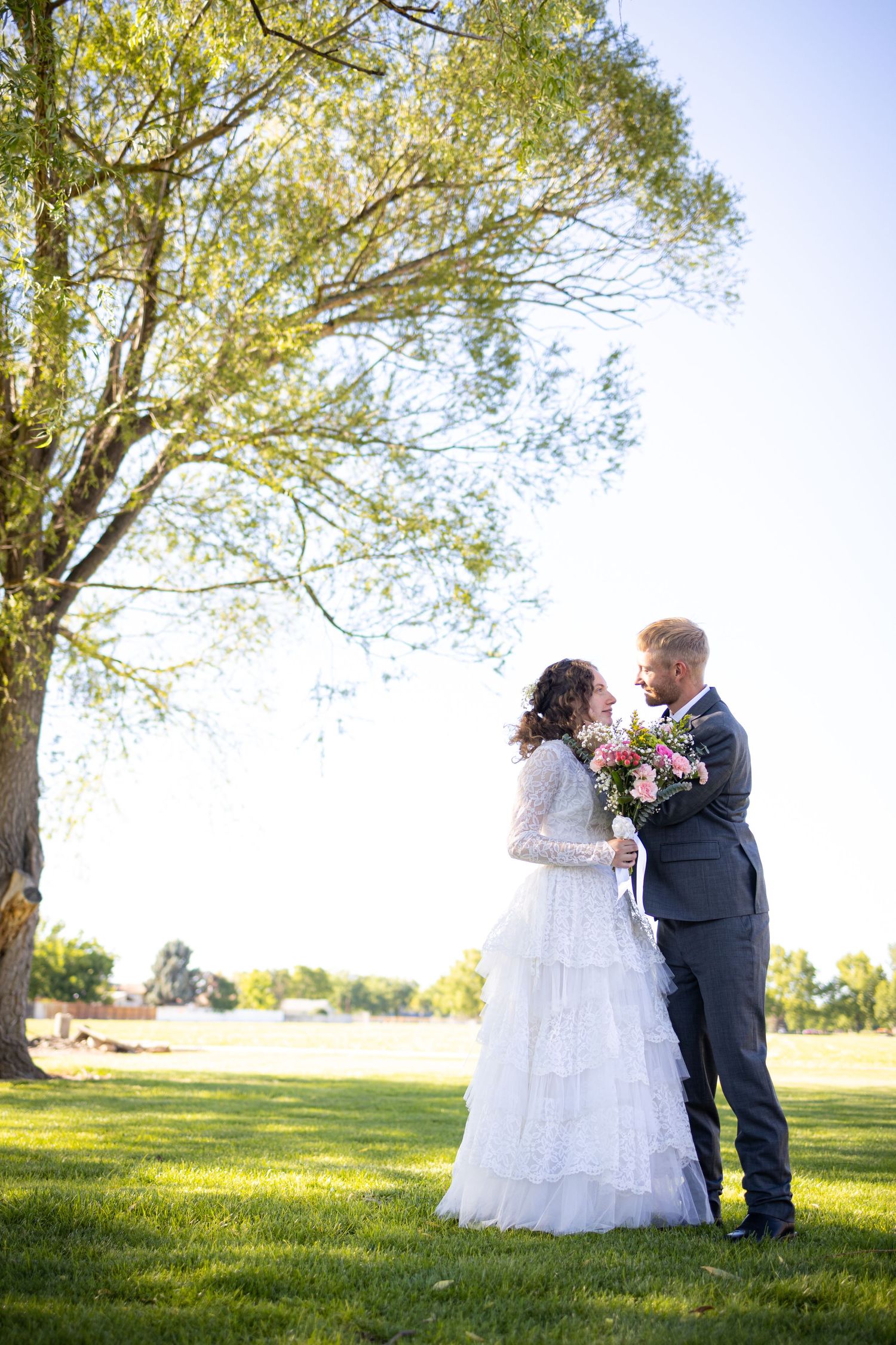 A scenic outdoor wedding portrait under a large tree in a sunny meadow.