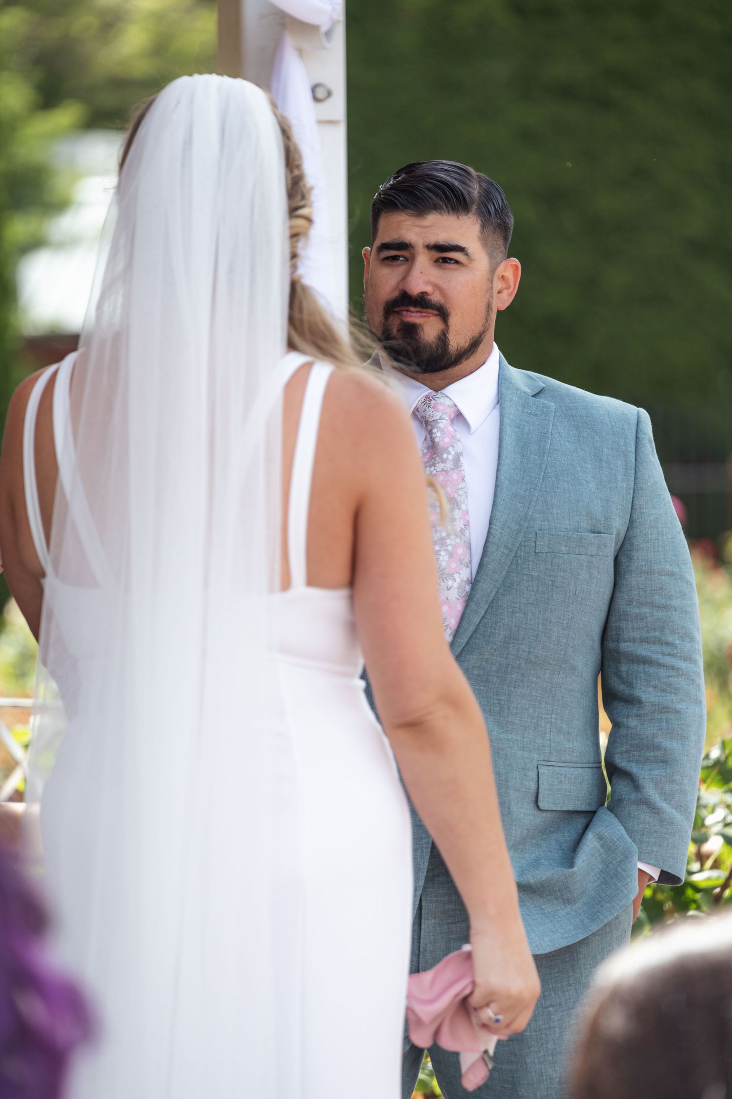 Bride in white dress and veil stands with wedding party during outdoor ceremony.