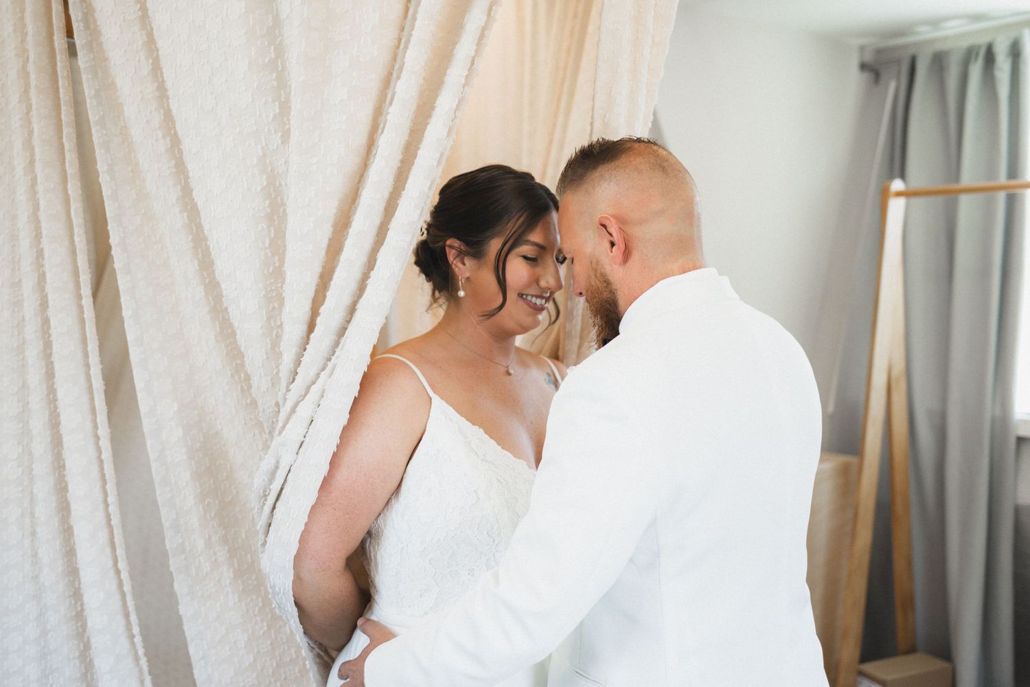 Romantic wedding portrait of couple in white sharing an intimate moment against light curtains.