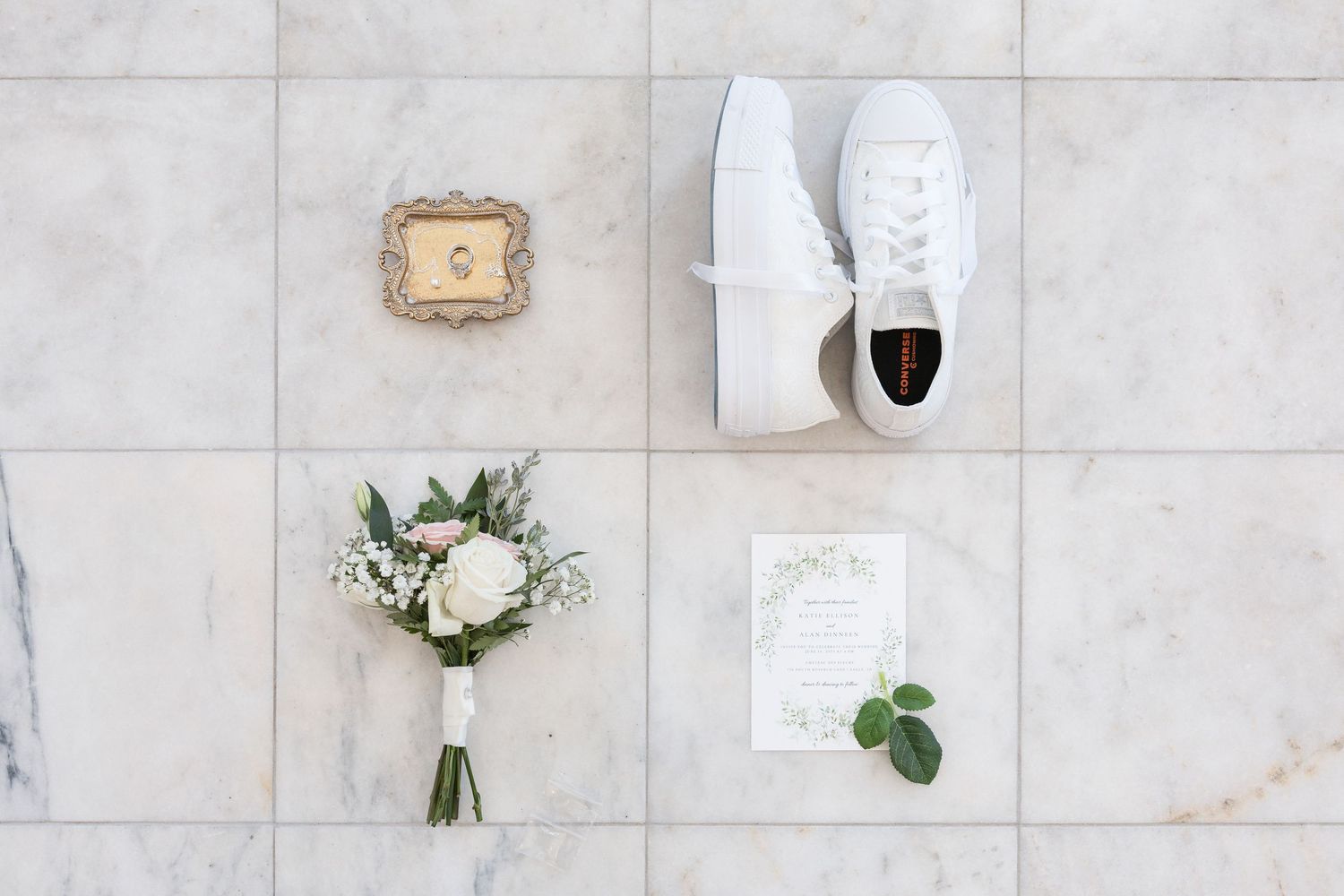 Wedding details including white shoes, invitations, and floral arrangements displayed on marble tile flooring.
