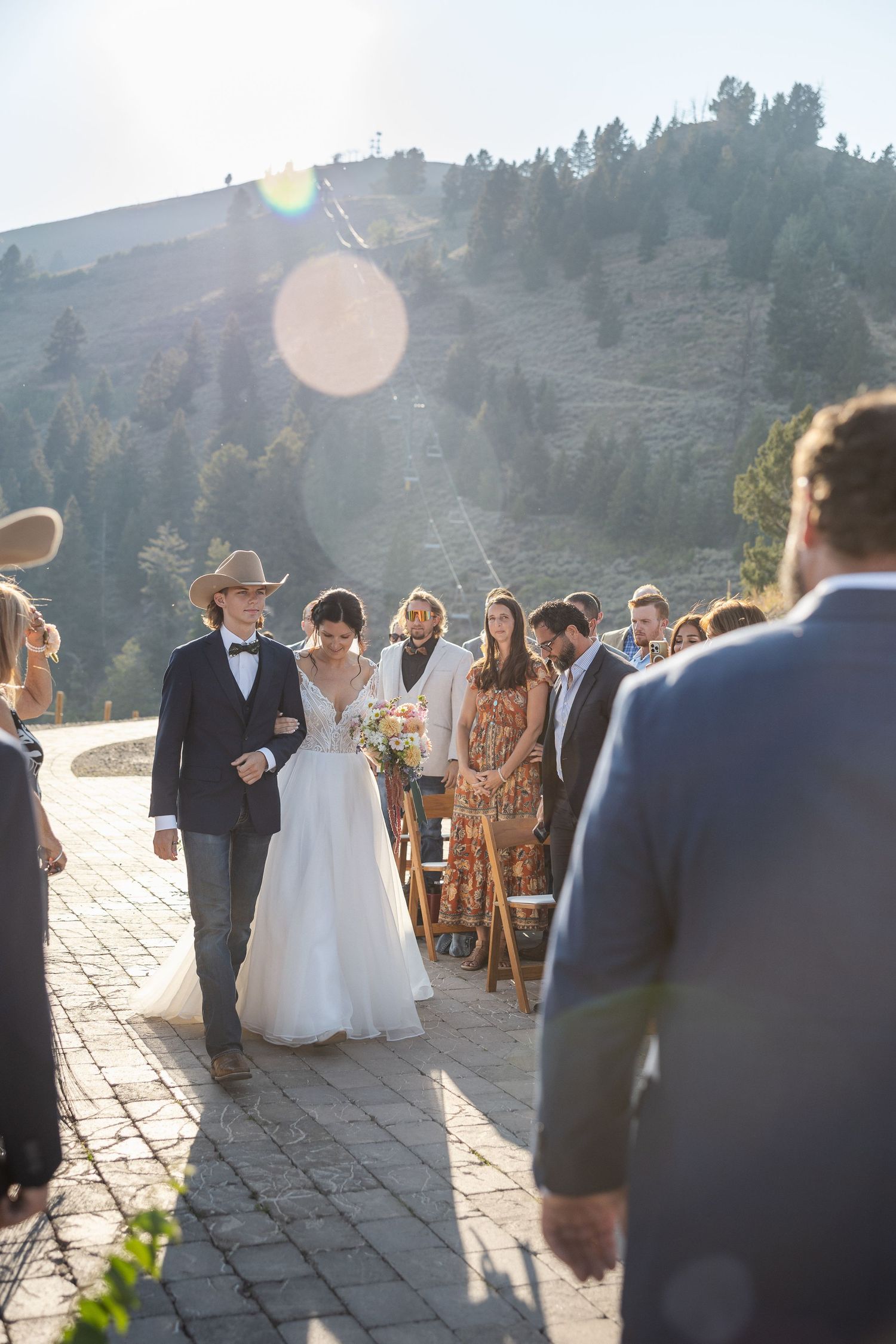 Outdoor mountain wedding ceremony with guests seated on wooden chairs as sunlight creates lens flares.