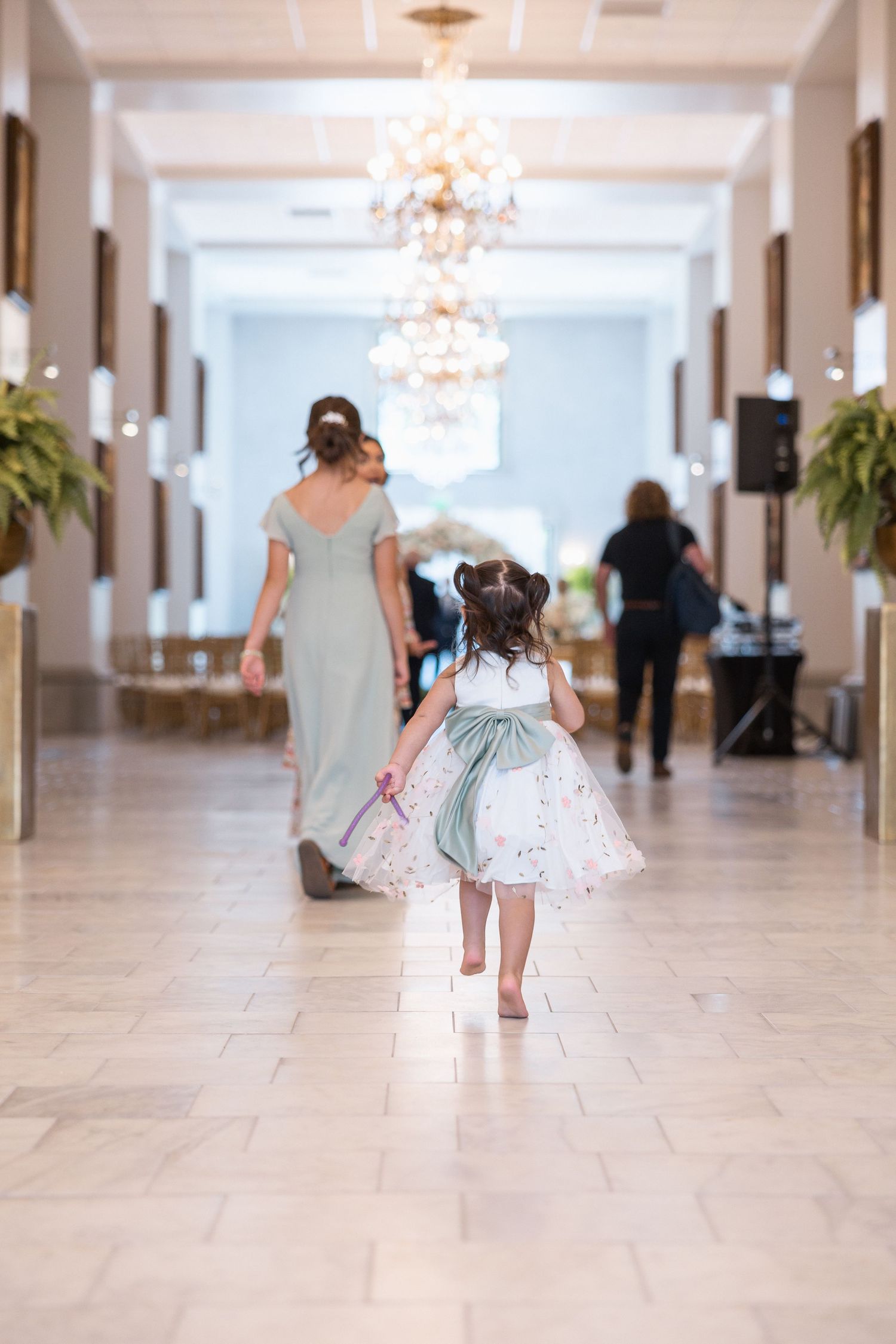 A little girl in a white dress runs down an elegant hallway with chandeliers.