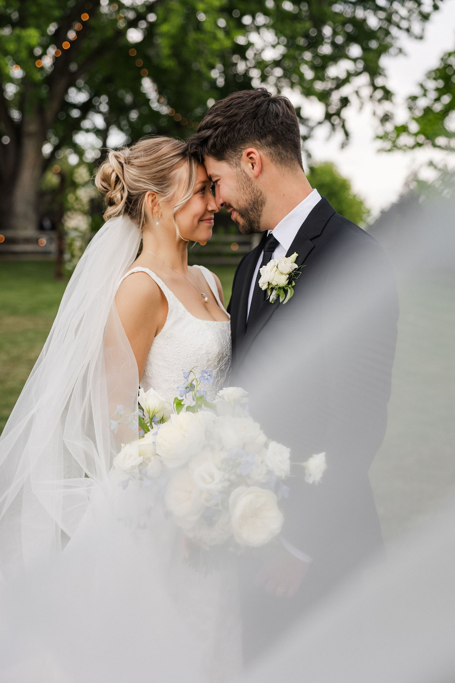 Bride and groom touching foreheads romantically outdoors, veil flowing in foreground.