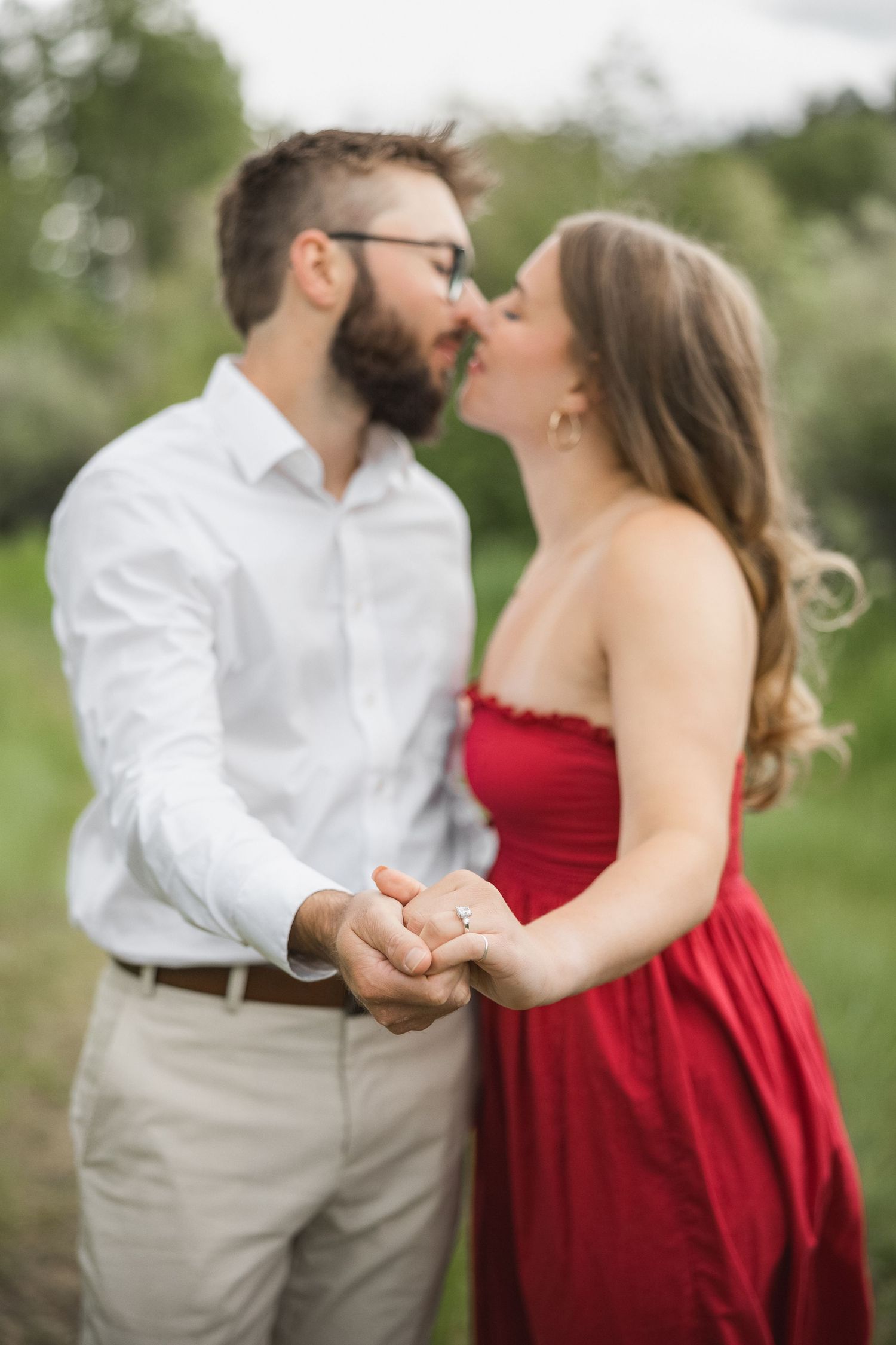 A couple shares a romantic kiss outdoors, with her in a red dress and him in a white shirt and khakis.