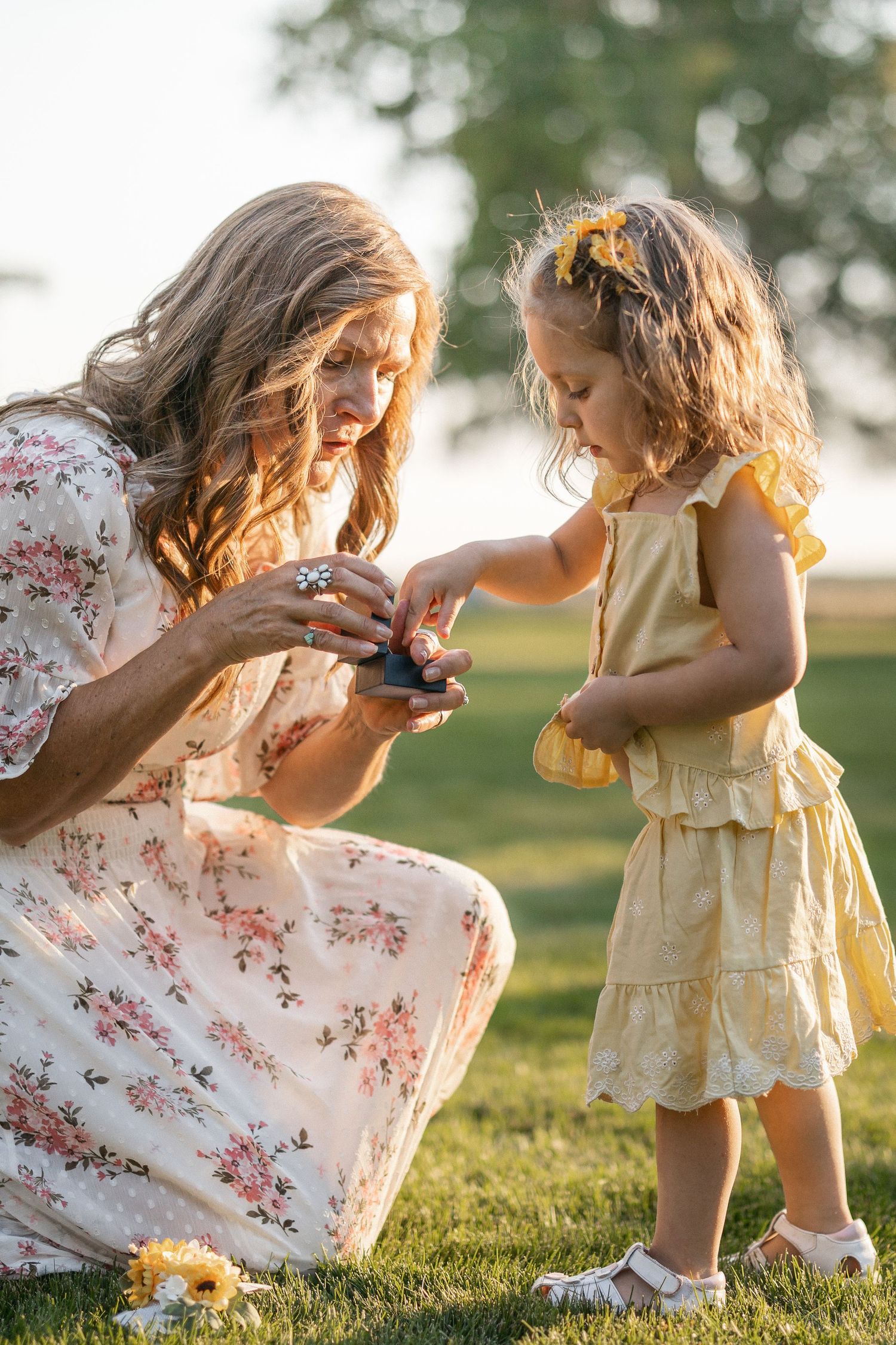 A tender outdoor moment between two people in floral and yellow dresses against a sunlit grassy backdrop.