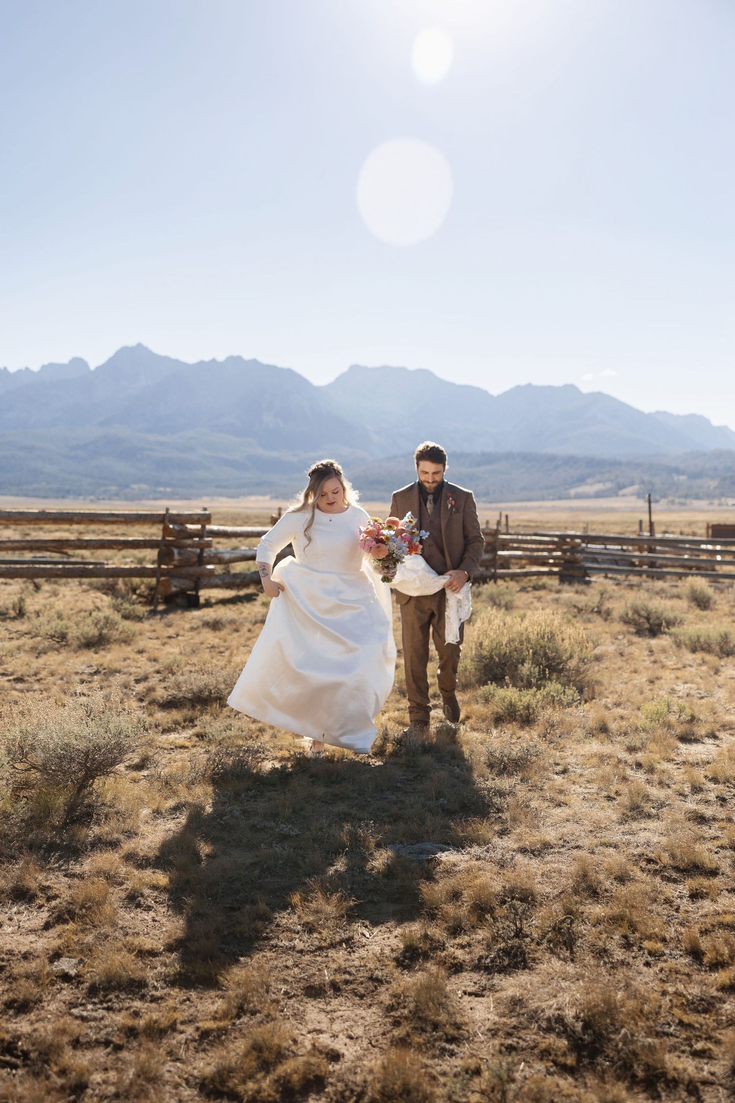 A couple celebrates their wedding day in a rustic field with mountain peaks in the background on a sunny afternoon.